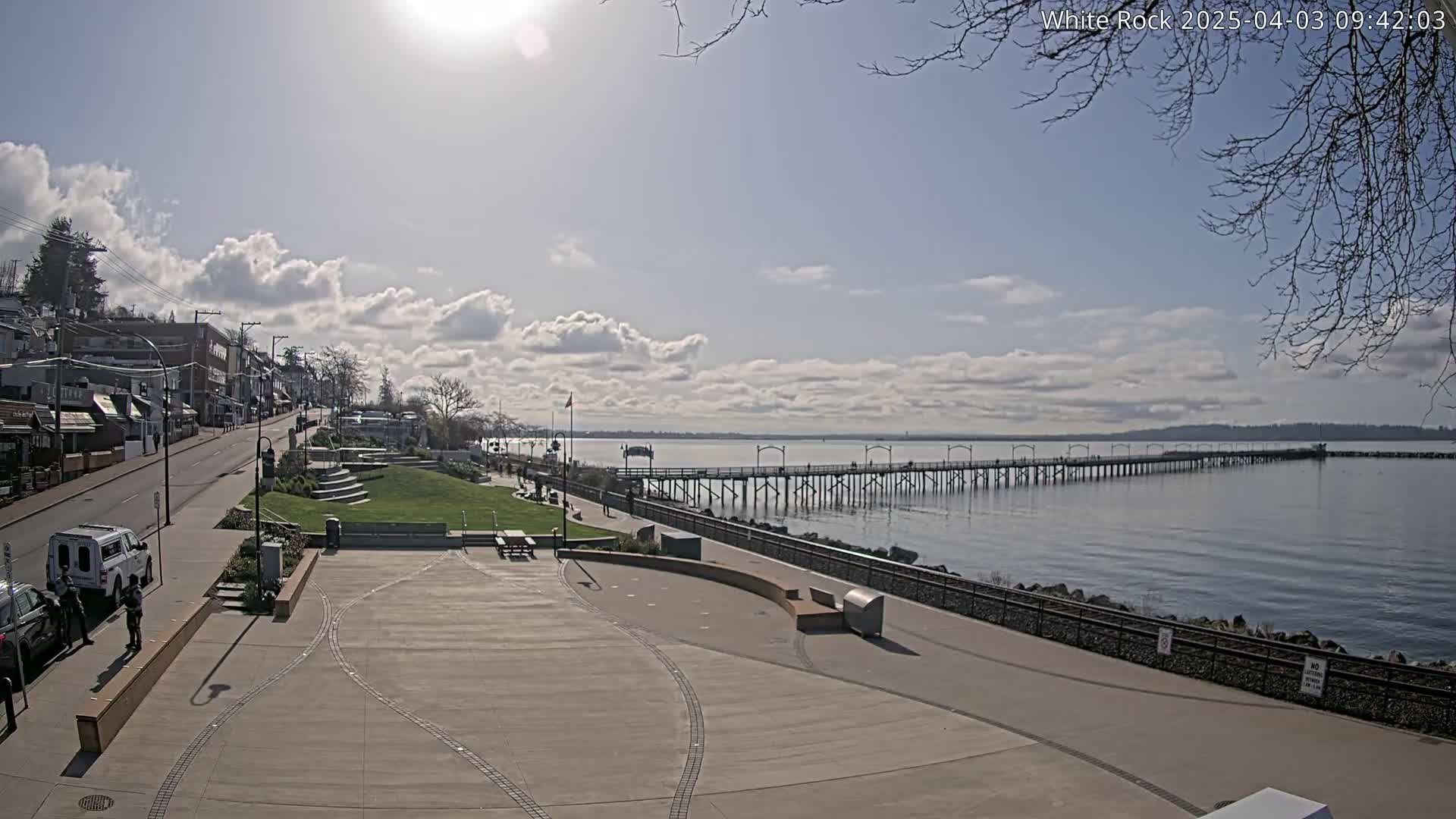 A sunny day with partly cloudy skies shows a waterfront town with a long pier extending into a calm bay, a paved plaza, and a street lined with buildings.