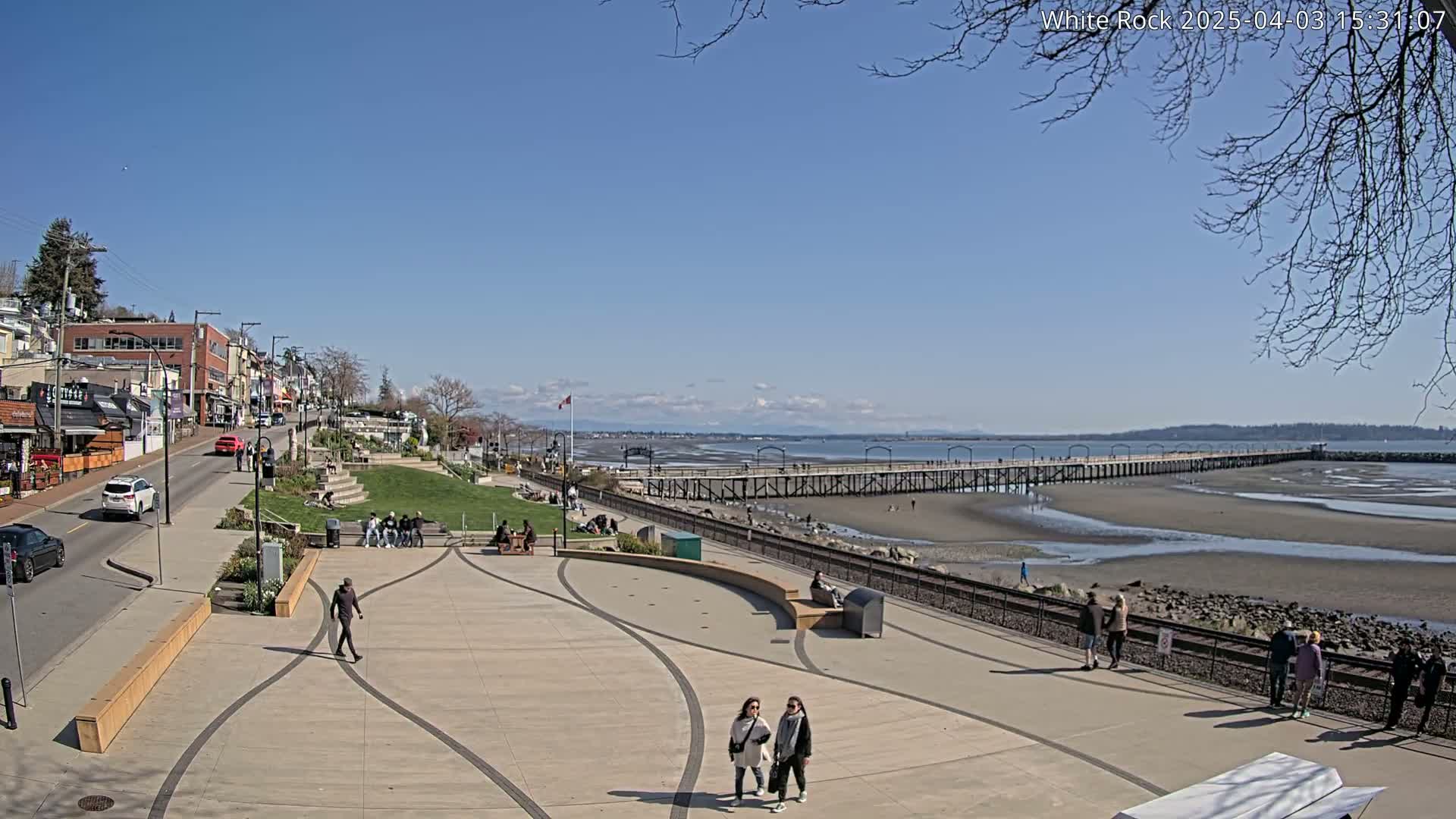 A paved waterfront plaza with a long pier extending into a mostly exposed sandy bay under a clear blue sky.