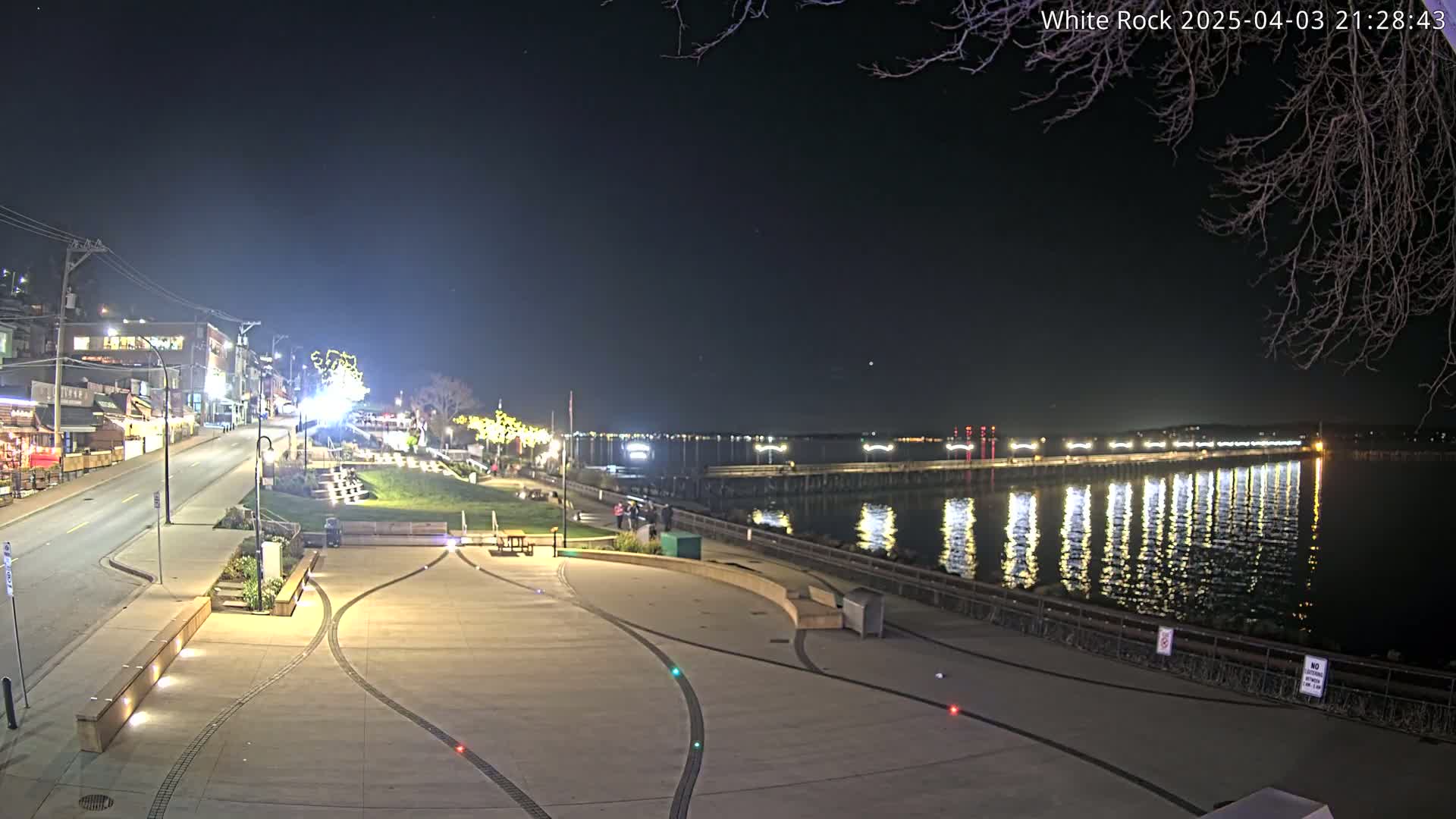 A nighttime view of a waterfront town square, with a long pier extending into calm, light-reflecting water under a clear, dark sky.