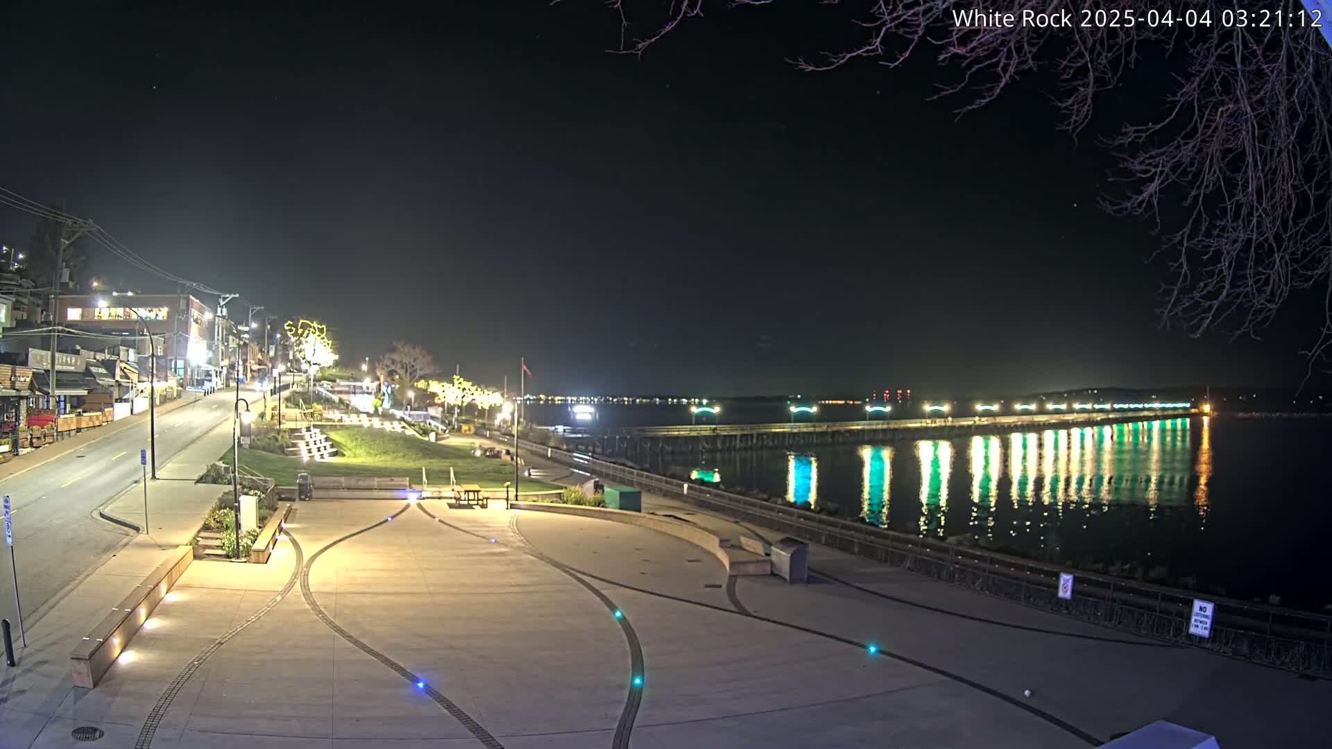 A nighttime view of a waterfront plaza with a long pier extending into the calm water, illuminated by lights reflecting on the surface.