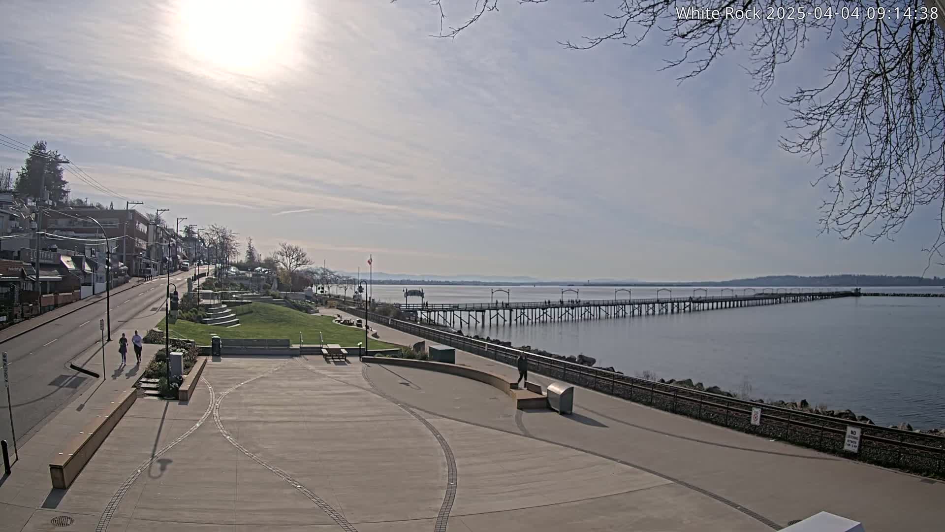A sunny day reveals a seaside town with a long pier extending into calm water, a paved plaza in the foreground, and a street lined with buildings.