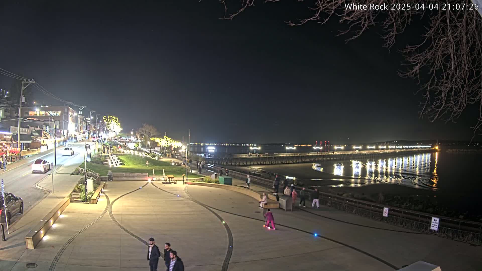A nighttime view of a waterfront town with a paved plaza, some cars, pedestrians, and a long pier extending into calm water reflecting lights.