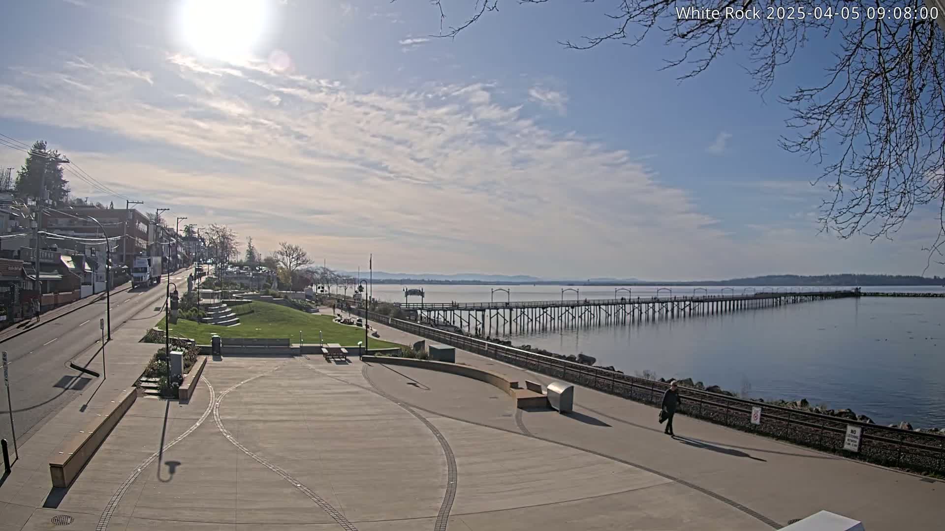 A long pier extends into a calm body of water under a partly sunny sky, overlooking a paved waterfront area with a street and buildings in the background.