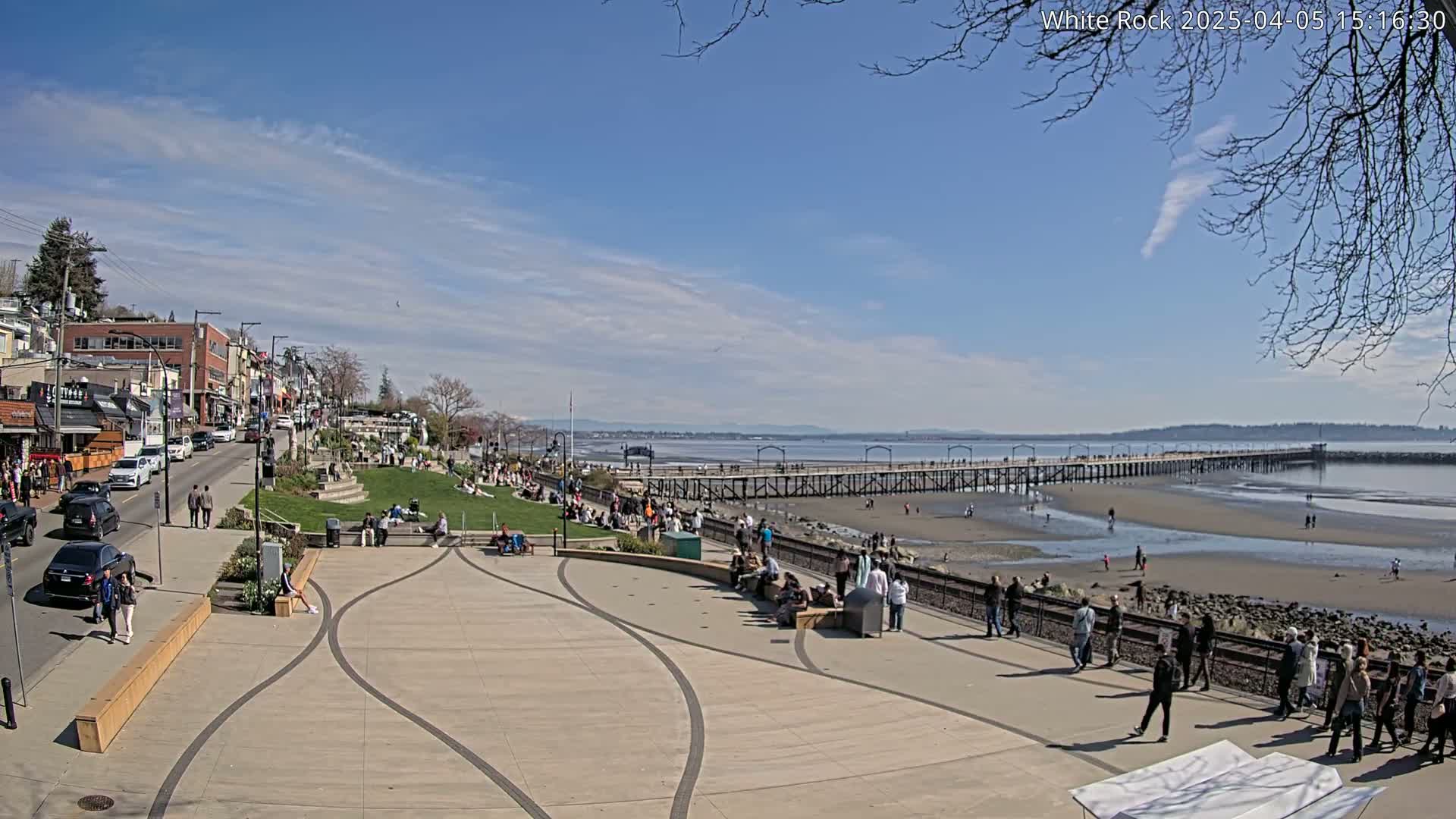 A sunny day at a waterfront plaza shows a long pier extending into a bay, with many people strolling along the plaza, the pier, and the beach.