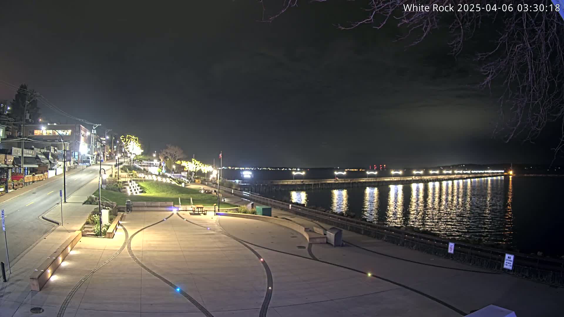 A nighttime view of a waterfront plaza with a paved area, benches, and low lighting, overlooking a calm body of water reflecting lights from a pier and distant shoreline under a dark, cloudy sky.