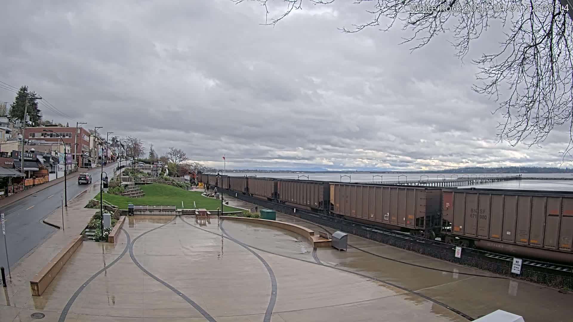 A wet, paved plaza area with a long freight train parked beside a calm body of water under an overcast sky.