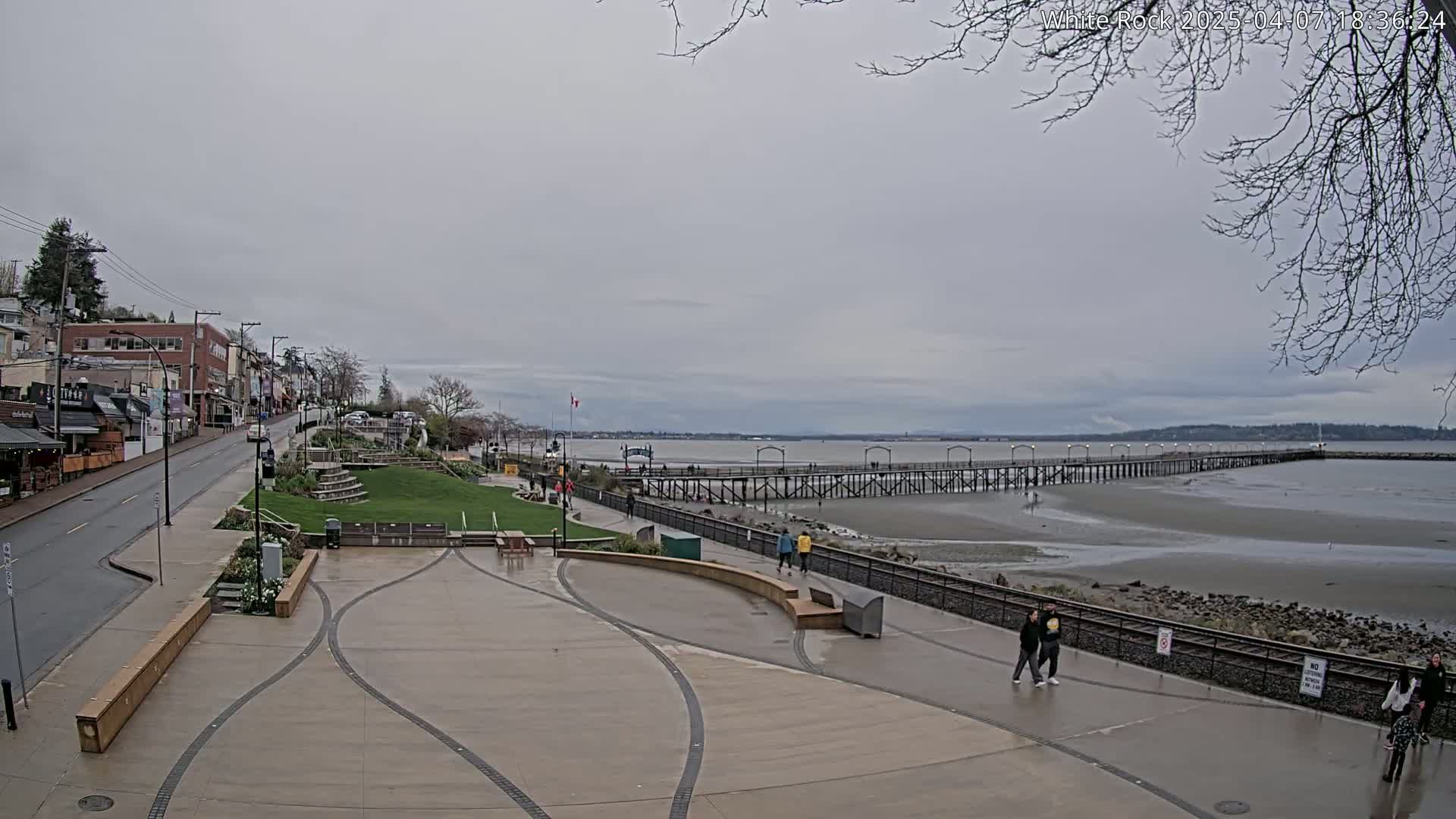 A seaside town on an overcast day features a long pier extending into shallow, exposed water, with people walking along a paved waterfront promenade.
