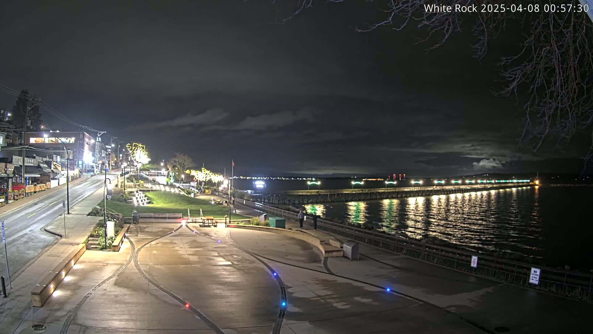 A nighttime view of a waterfront area with a long pier extending into the calm water, under a partly cloudy sky, and a paved walkway leading to it from a street lined with buildings.