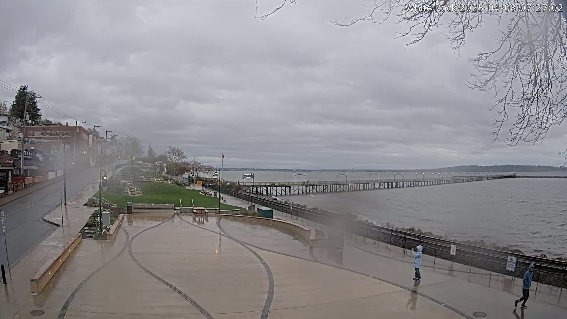 A wet plaza and walkway lead to a long pier extending into a gray, choppy sea under a cloudy sky.