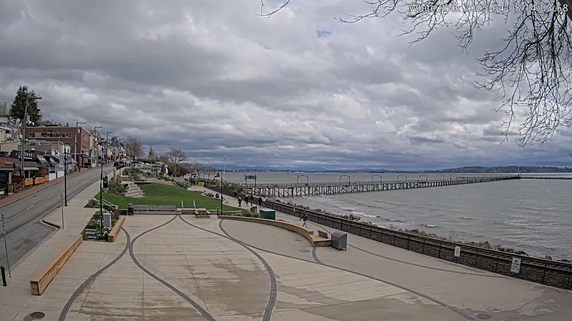 A paved waterfront plaza with a long pier extending into a calm, gray sea under a cloudy sky.