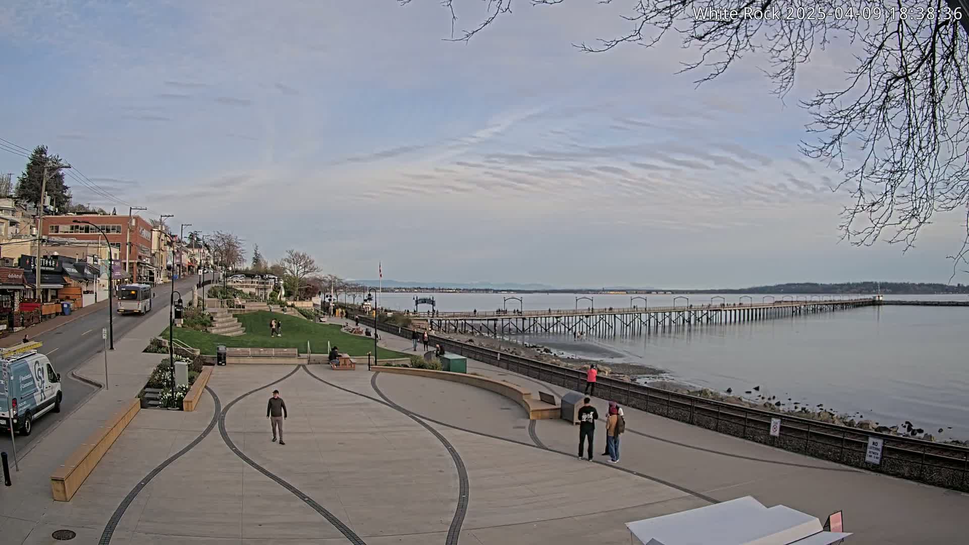 A long pier extends into a calm body of water under a partly cloudy sky, with a paved waterfront area featuring a plaza, buildings, and a few pedestrians.
