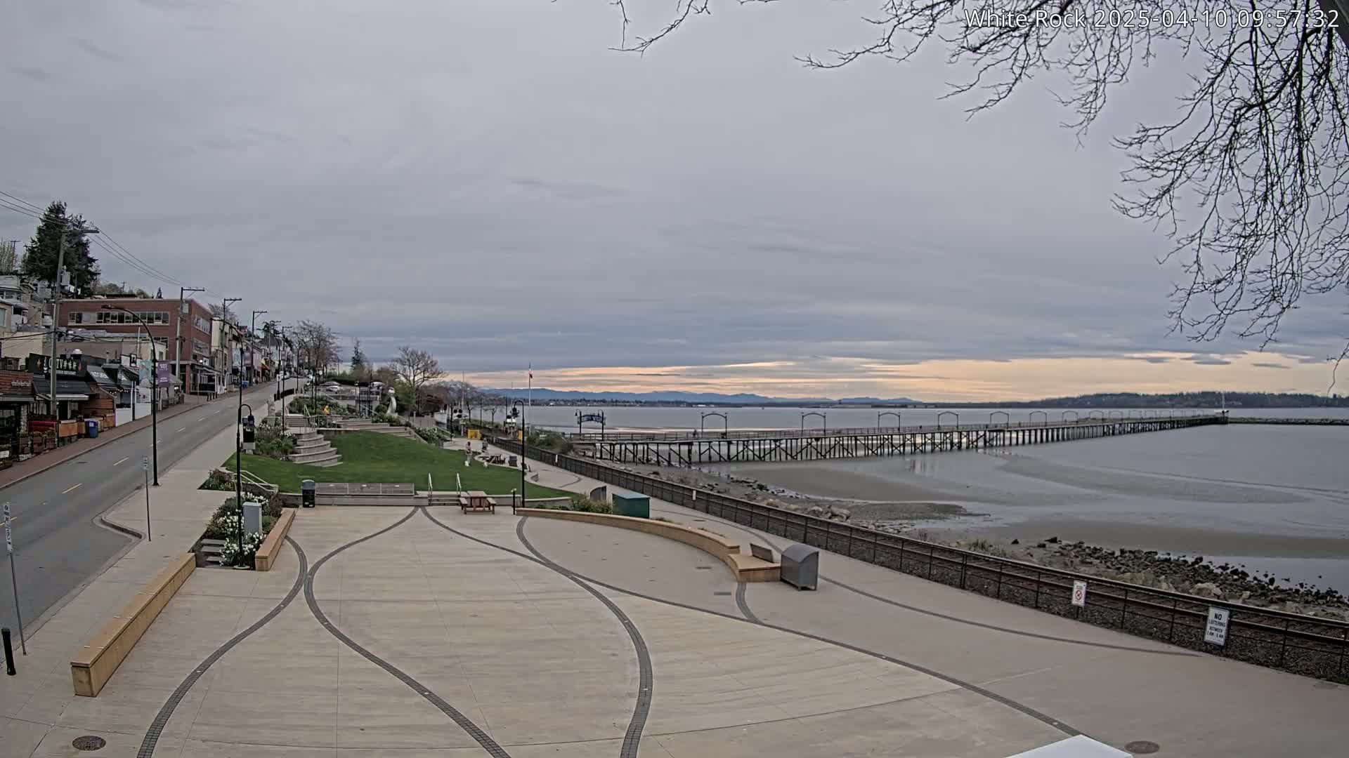 A paved waterfront plaza with a long pier extending into a calm, overcast bay, backed by a street and buildings.