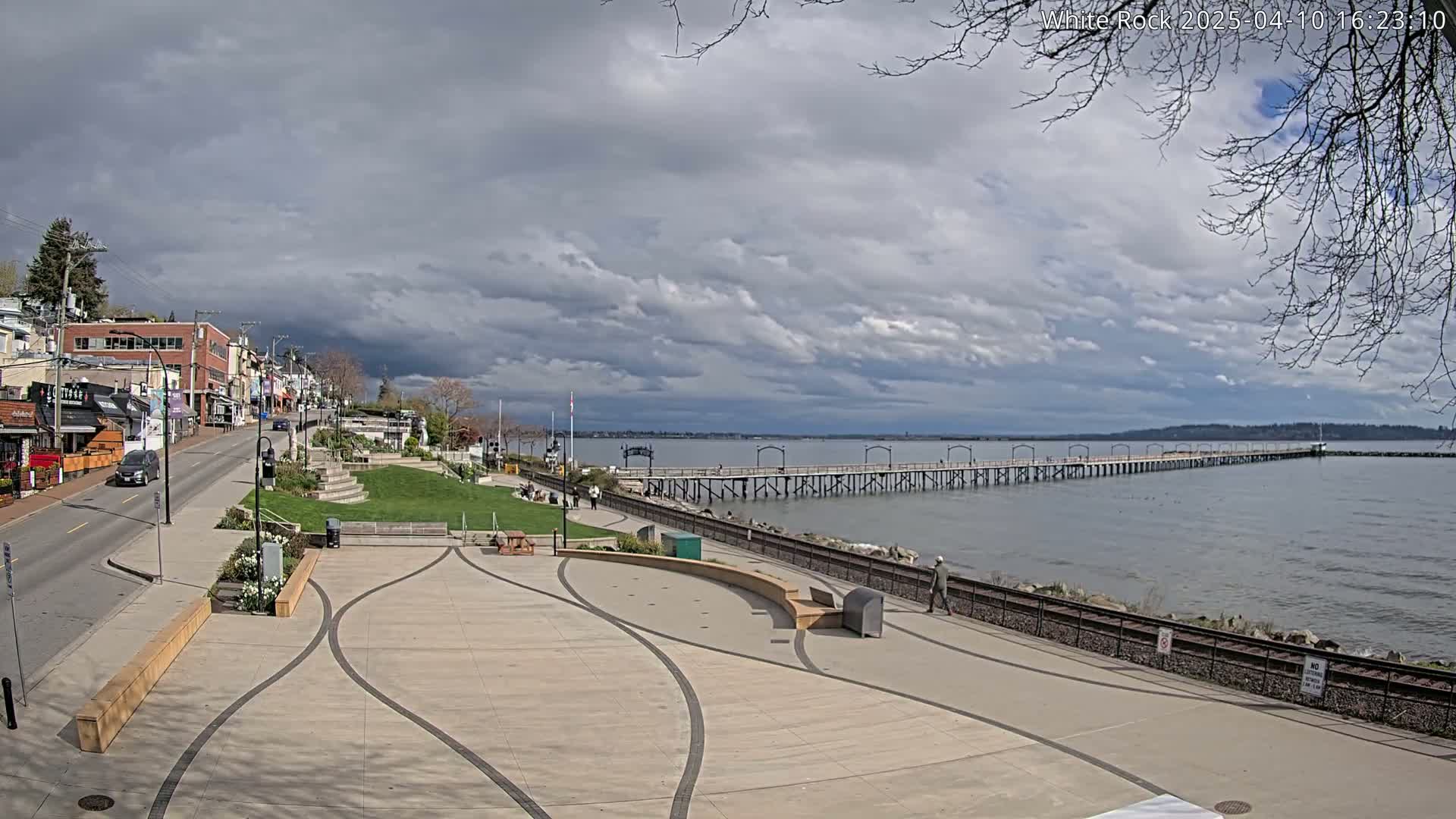 A long pier extends into a calm body of water under a partly cloudy sky, with a paved waterfront area and shops visible on the left.