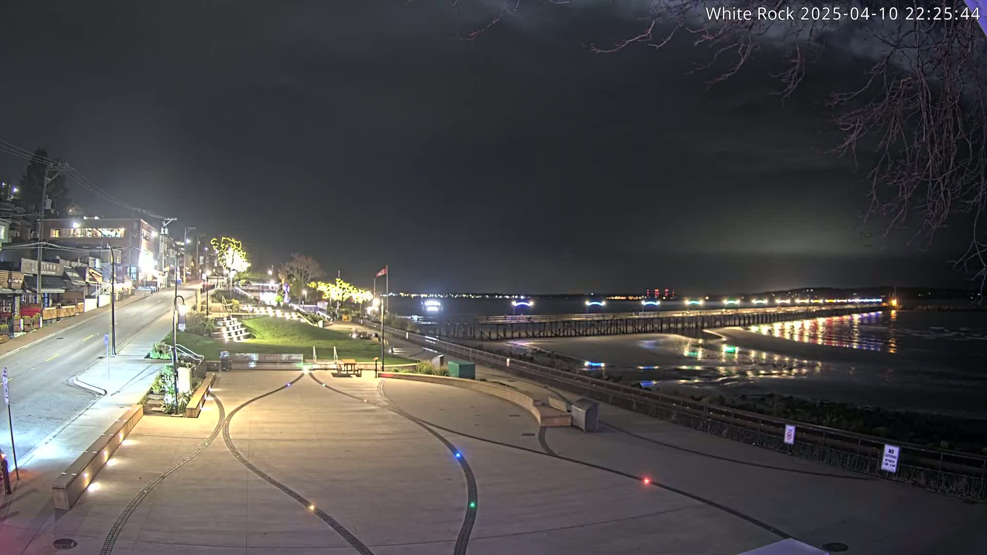 A nighttime view of a waterfront area shows a paved plaza, a street with buildings, and a pier extending into a calm, dark body of water reflecting lights.