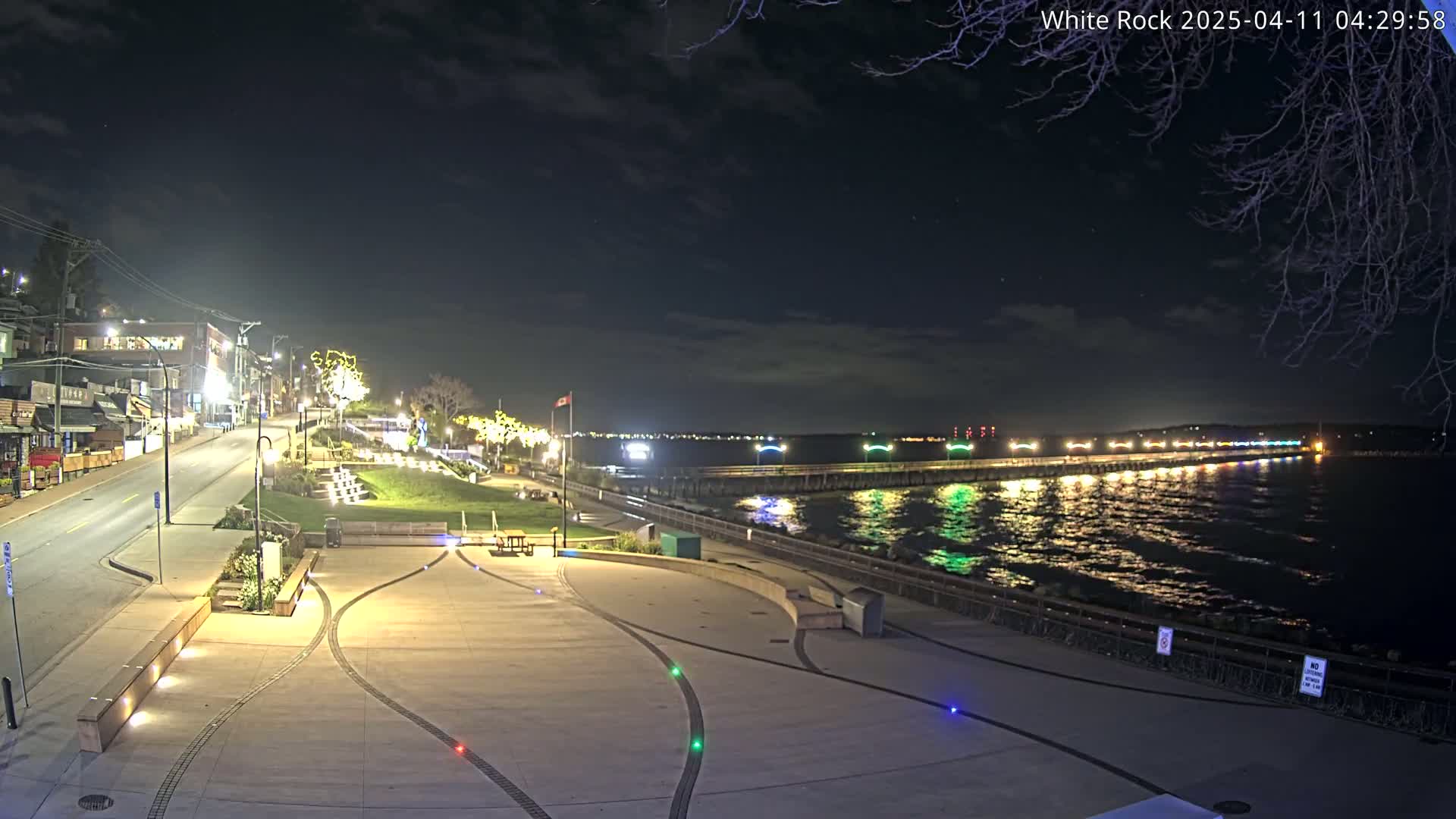 A nighttime view of a waterfront plaza with a long pier extending into a calm, dark-water body, illuminated by colorful lights along the pier and on the plaza.