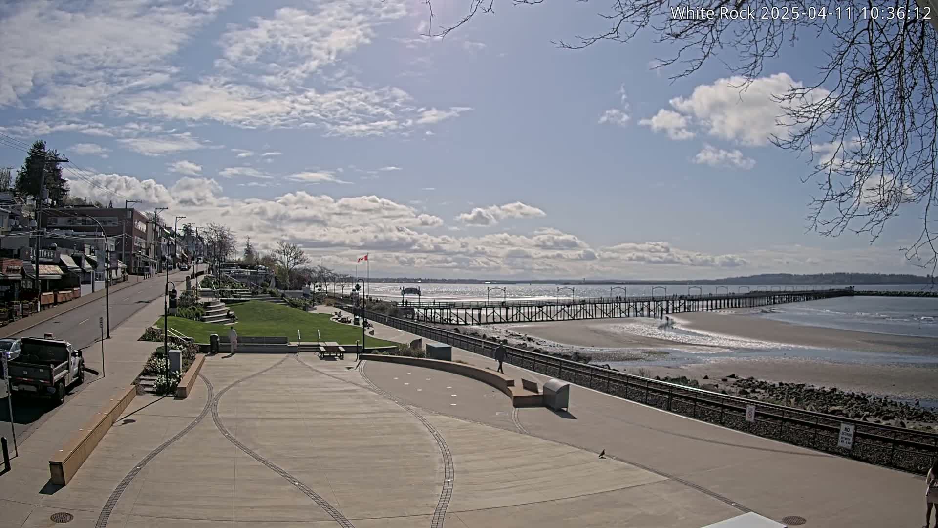 A paved waterfront plaza overlooks a sandy beach, a long pier, and calm ocean water under a partly sunny sky with scattered clouds.