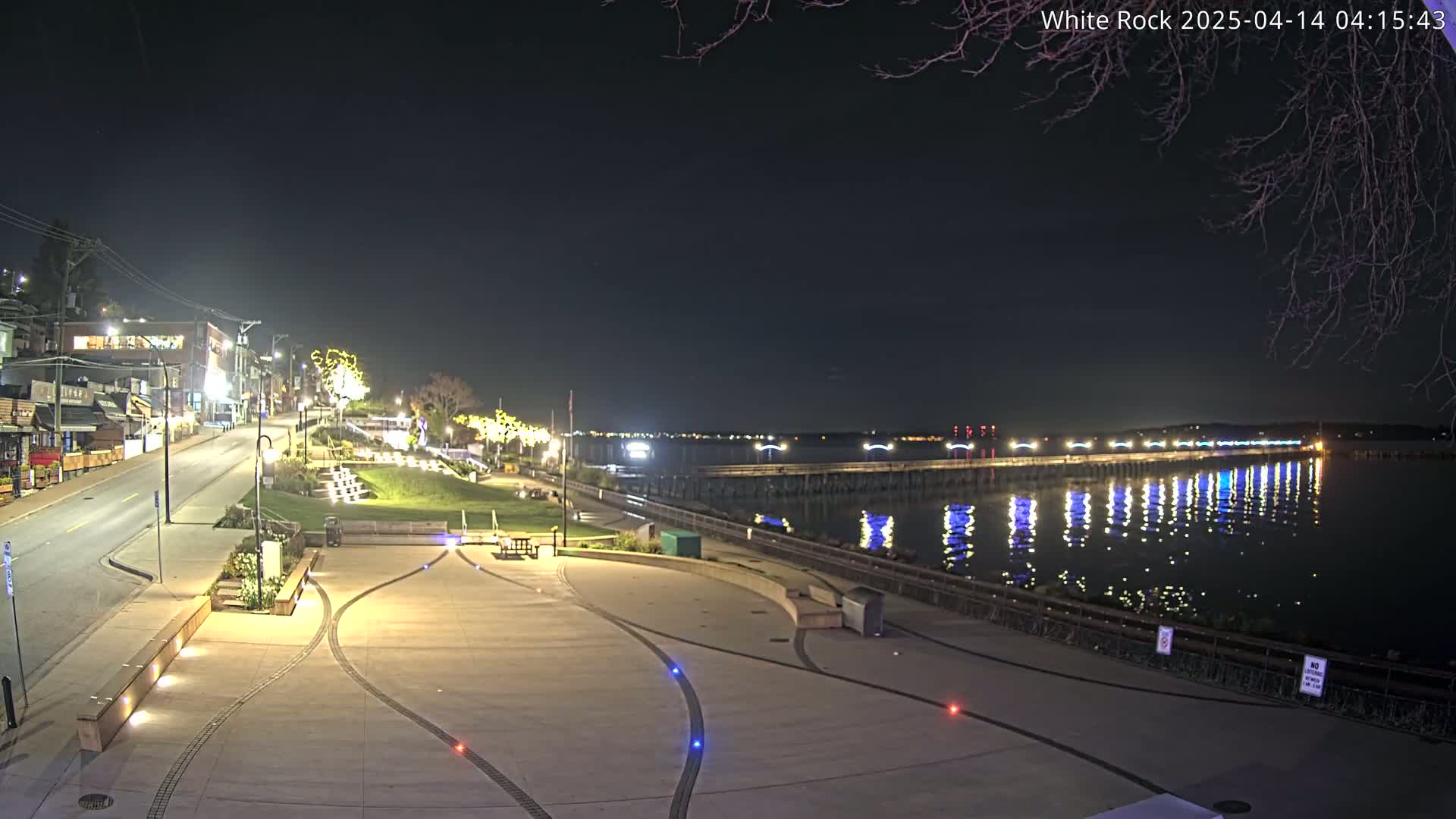 A nighttime view of a waterfront plaza with a long pier extending into calm water, illuminated by lights, under a clear, dark sky.