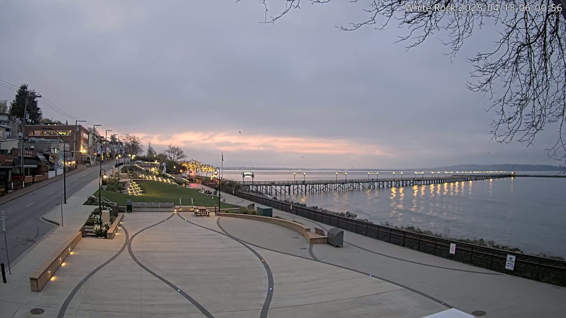 A long pier extends into a calm, twilight ocean beside a paved waterfront area with buildings and landscaping.