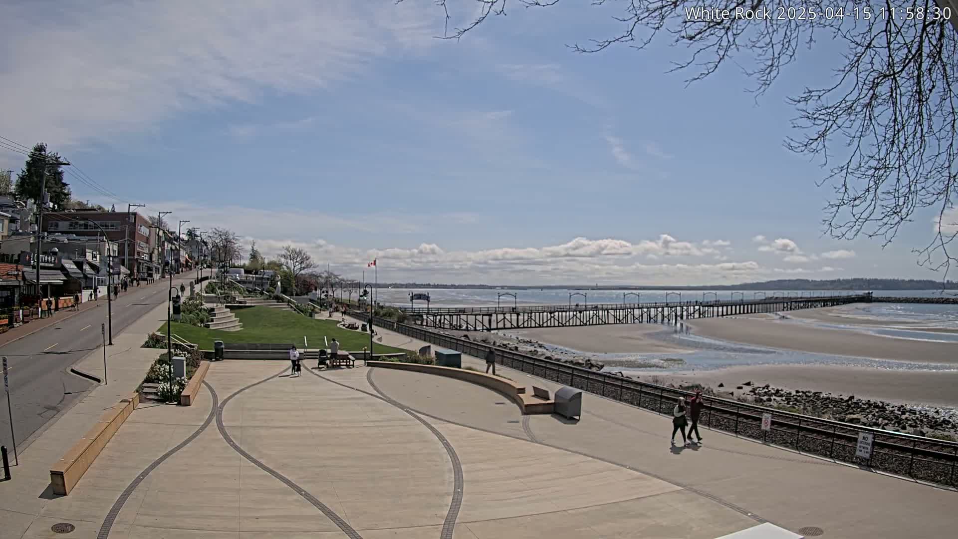 A long pier extends over a sandy beach under a partly cloudy blue sky, with a paved walkway and buildings lining the shore.