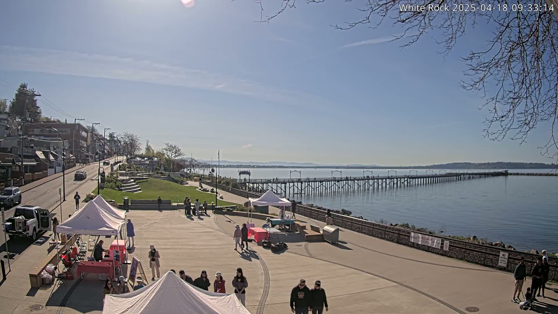 A sunny day reveals a waterfront plaza with several white tents, people strolling, a long pier extending into calm blue water, and a town visible in the background.