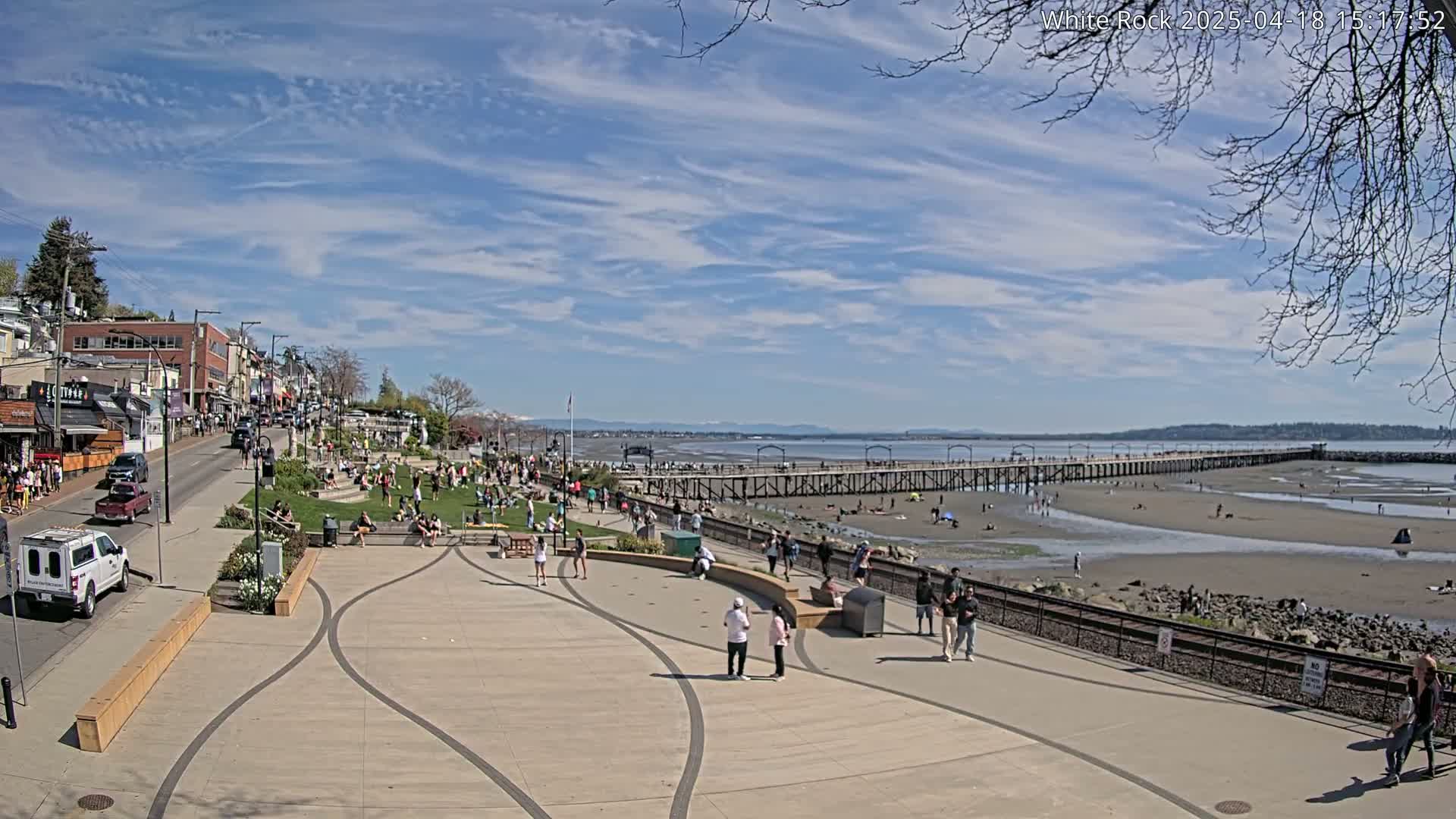 A sunny day shows a waterfront scene with many people enjoying a beach and pier, shops and buildings lining a street behind them.