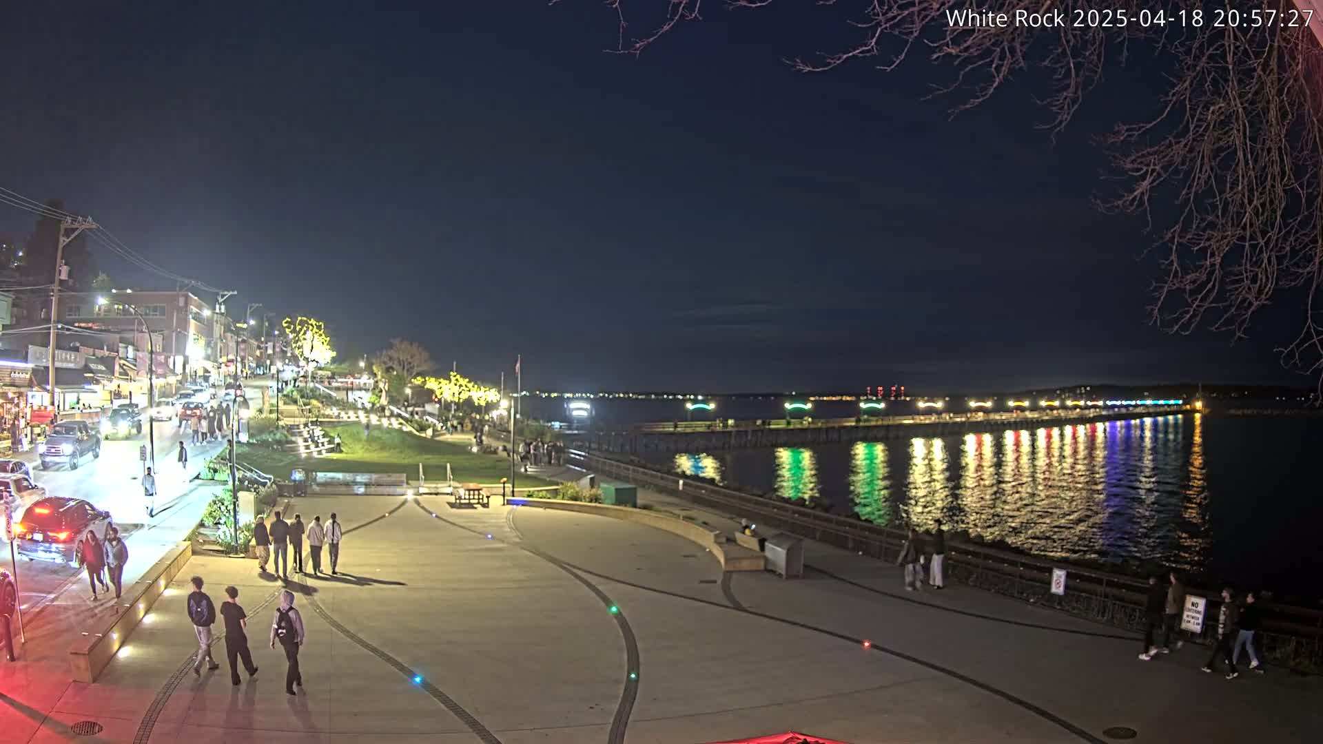 A nighttime waterfront scene shows a paved plaza with people walking, a street with cars, and a brightly lit pier extending into calm water reflecting the lights.