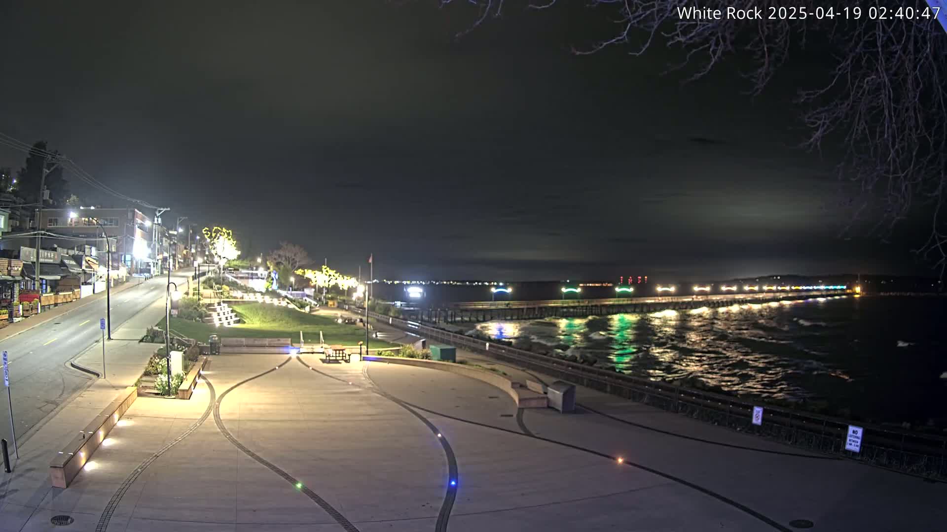 A nighttime view of a paved waterfront area with a long pier extending into a dark, wavy ocean under a mostly clear sky.