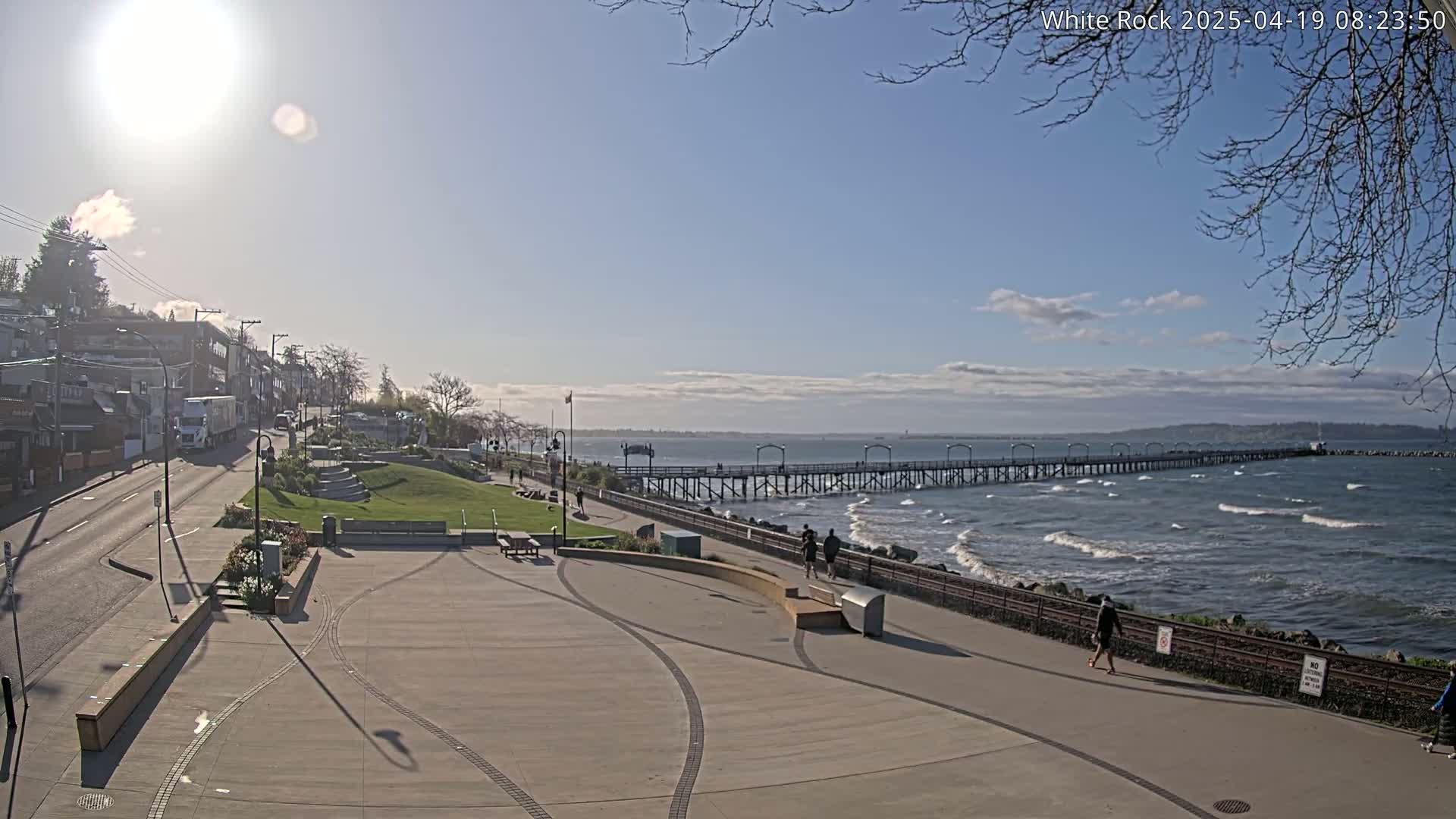 A sunny day reveals a seaside town with a long pier extending into a wavy ocean, a paved walkway, and a grassy park area with people strolling along the shore.