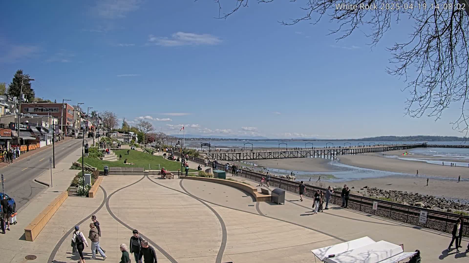 A sunny day at a waterfront town shows a paved plaza with people walking, a grassy area with people sitting, and a long pier extending into a sandy beach and calm water under a mostly clear sky.