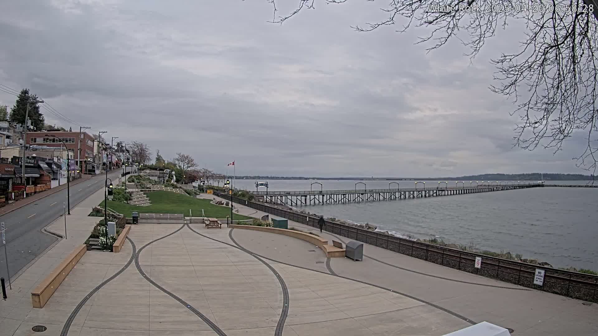 A long pier extends into a calm, grey-sky ocean, adjacent to a paved plaza and a street lined with buildings.