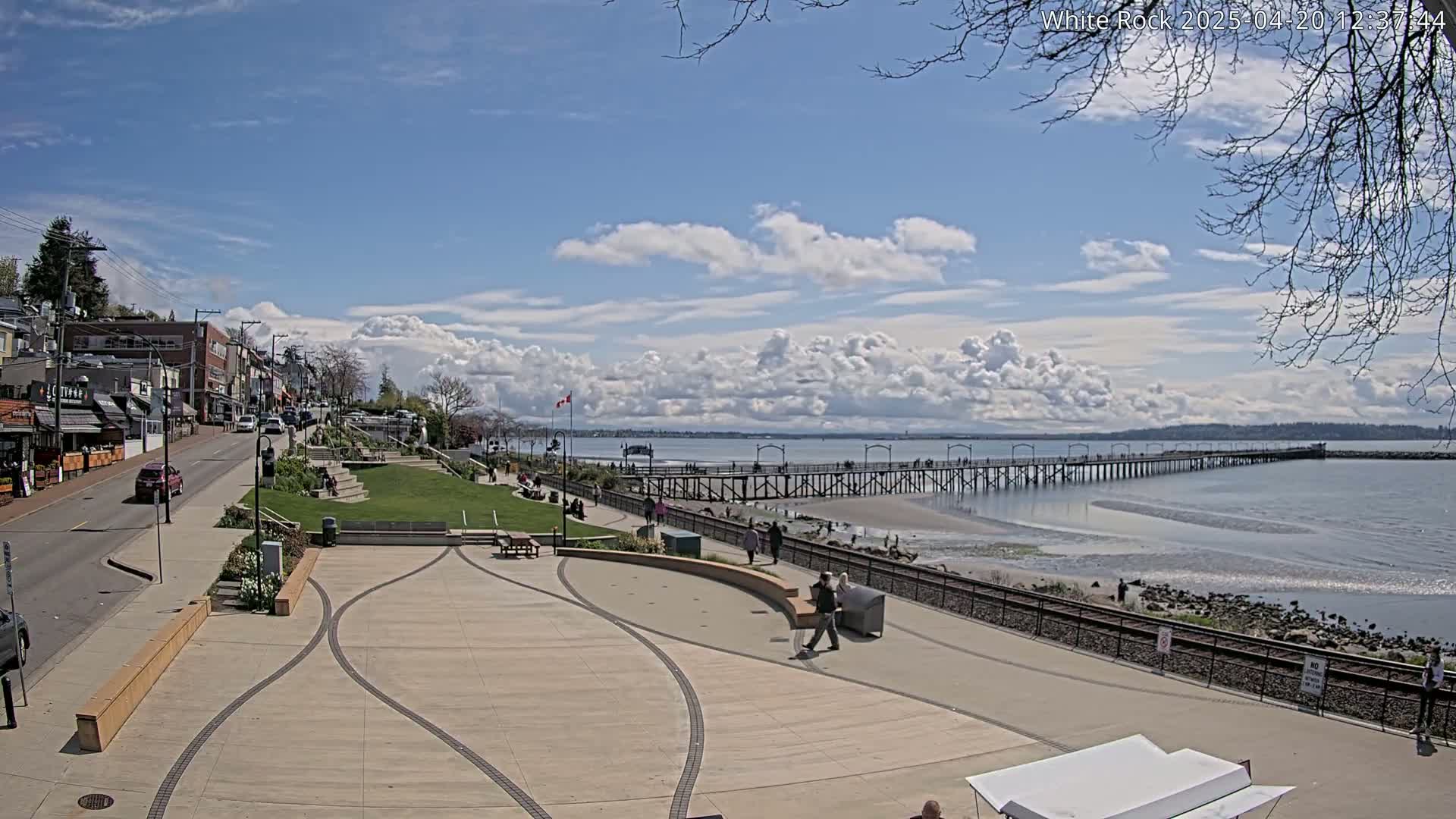 A paved waterfront plaza overlooks a long pier extending into a calm bay under a partly cloudy sky.