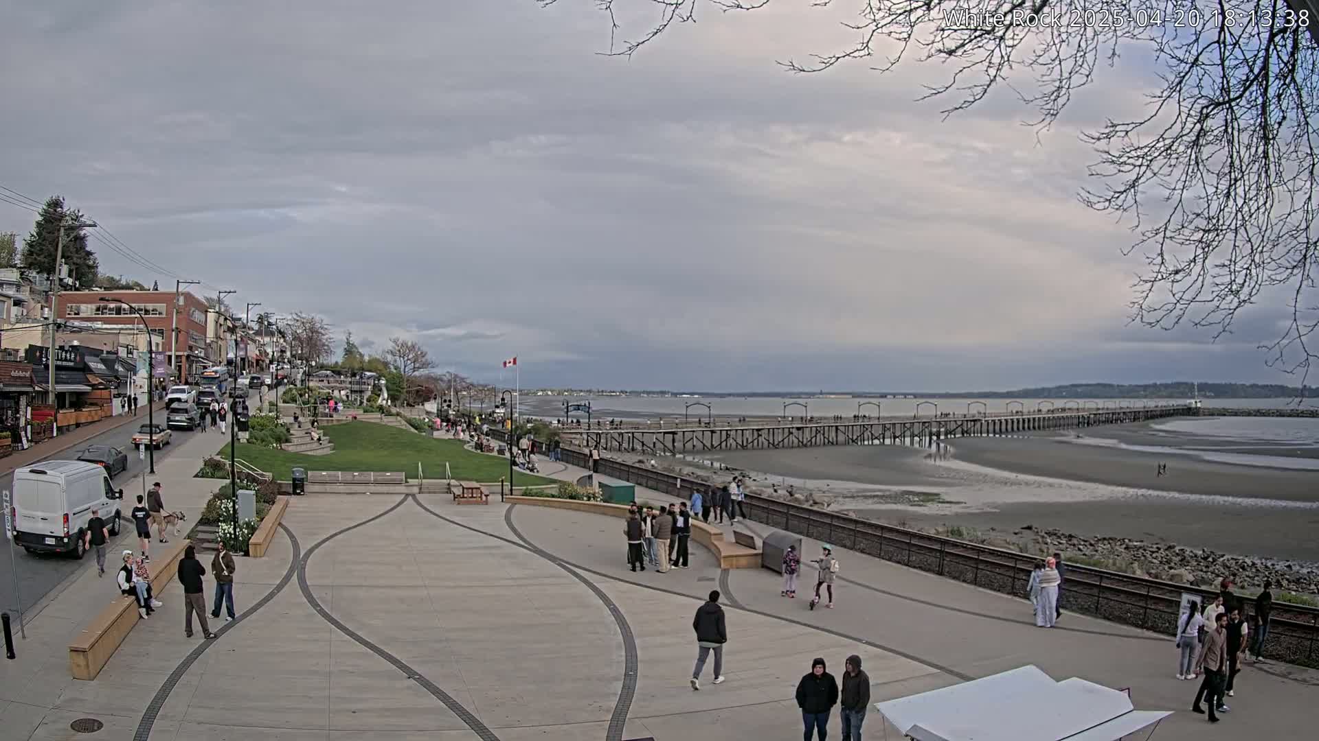 A paved waterfront plaza with people walking, shops and buildings along a street behind it, a long pier extending into a mostly exposed, sandy bay under a cloudy sky.