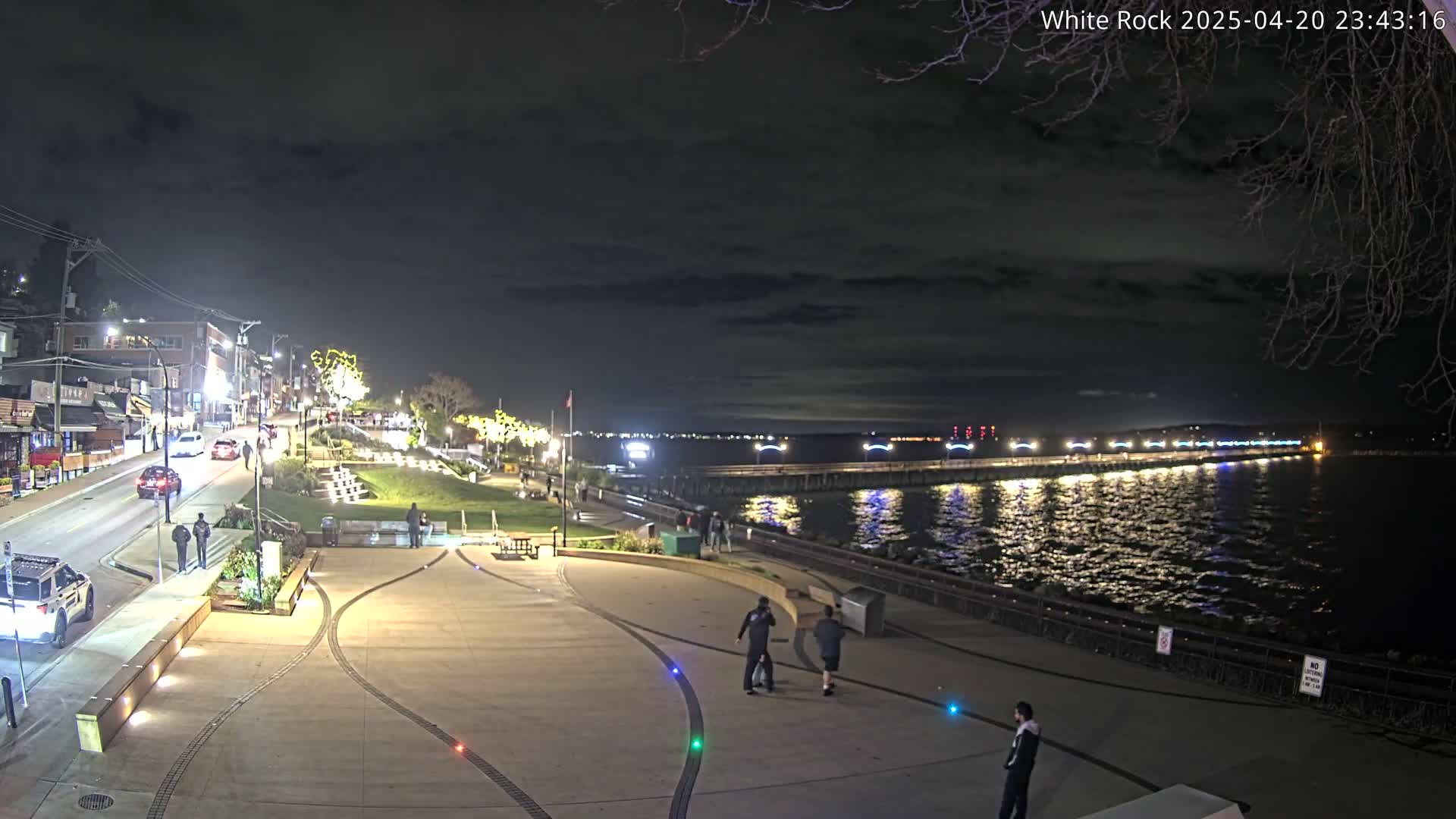 A nighttime view of a waterfront town with a long pier extending into a calm, dark body of water under a partly cloudy sky, featuring people walking along the shore and cars on a nearby road.