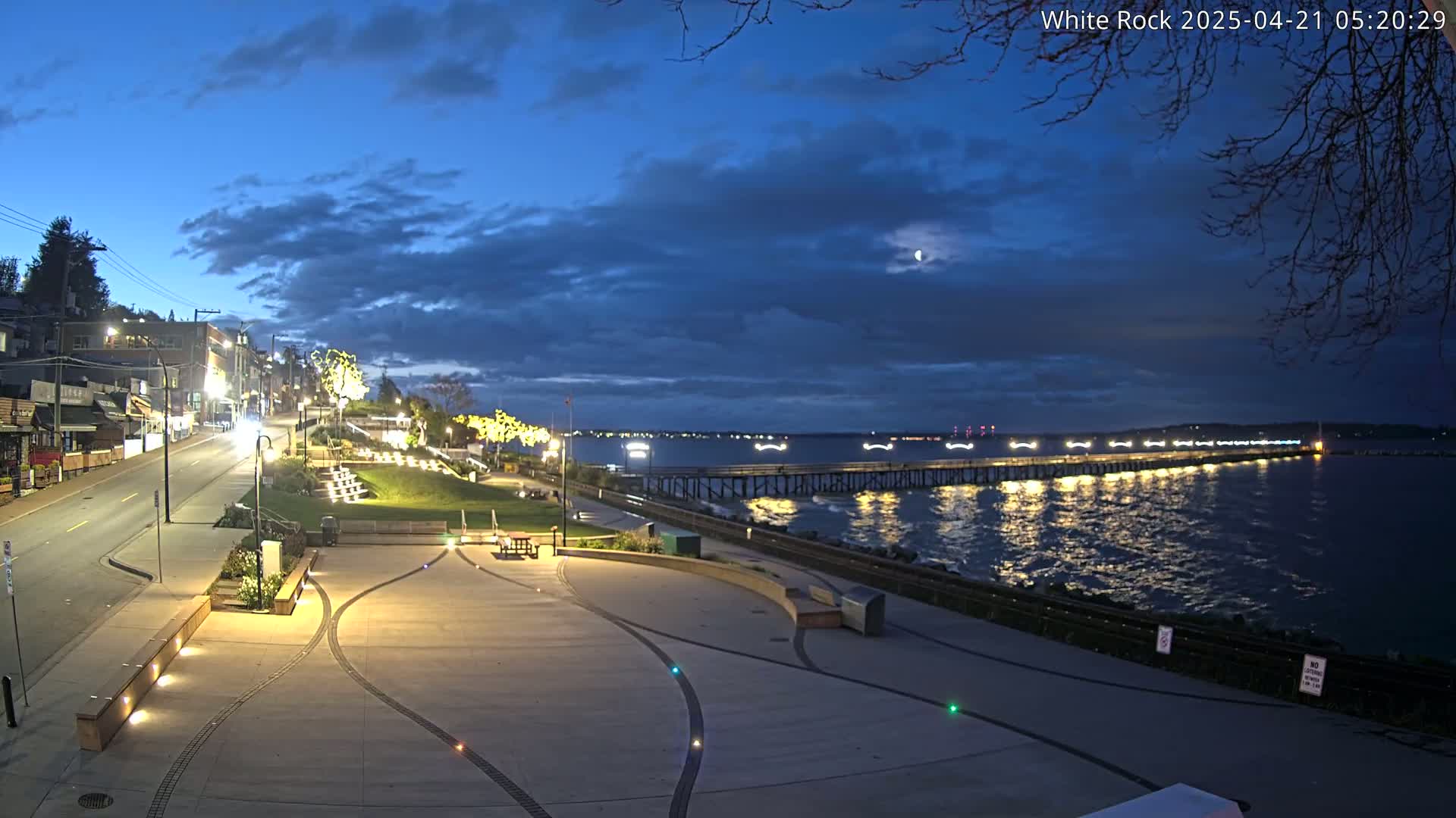 A nighttime scene shows a paved waterfront area with a long pier extending into a calm, dark blue body of water under a partly cloudy sky.