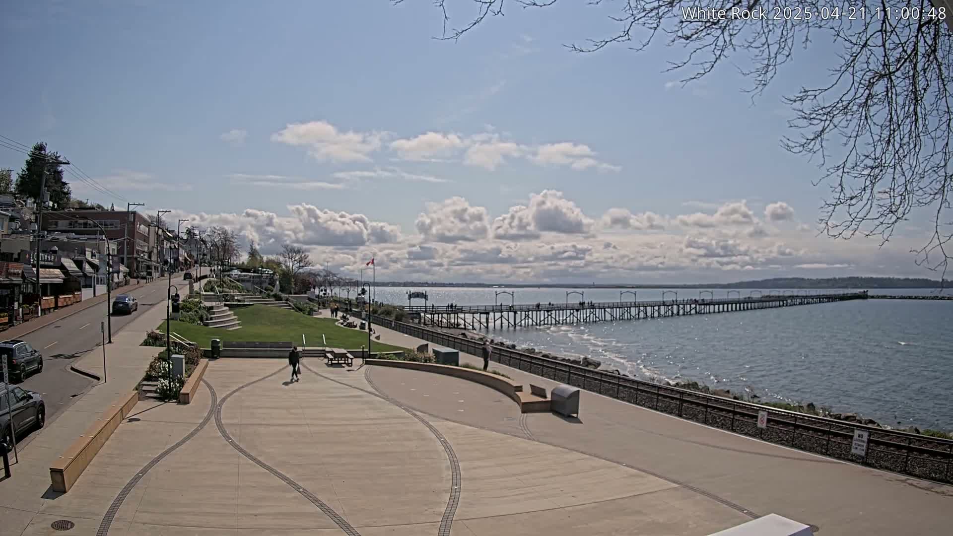 A seaside town square with a long pier extending into a calm ocean under a partly cloudy sky.