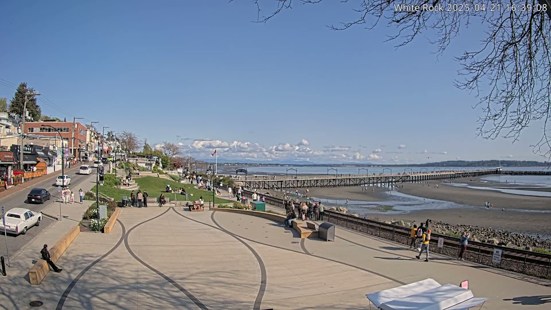 A sunny day shows a waterfront plaza with people strolling, sitting, and enjoying the view of a long pier extending into a wide, sandy beach with low tide.