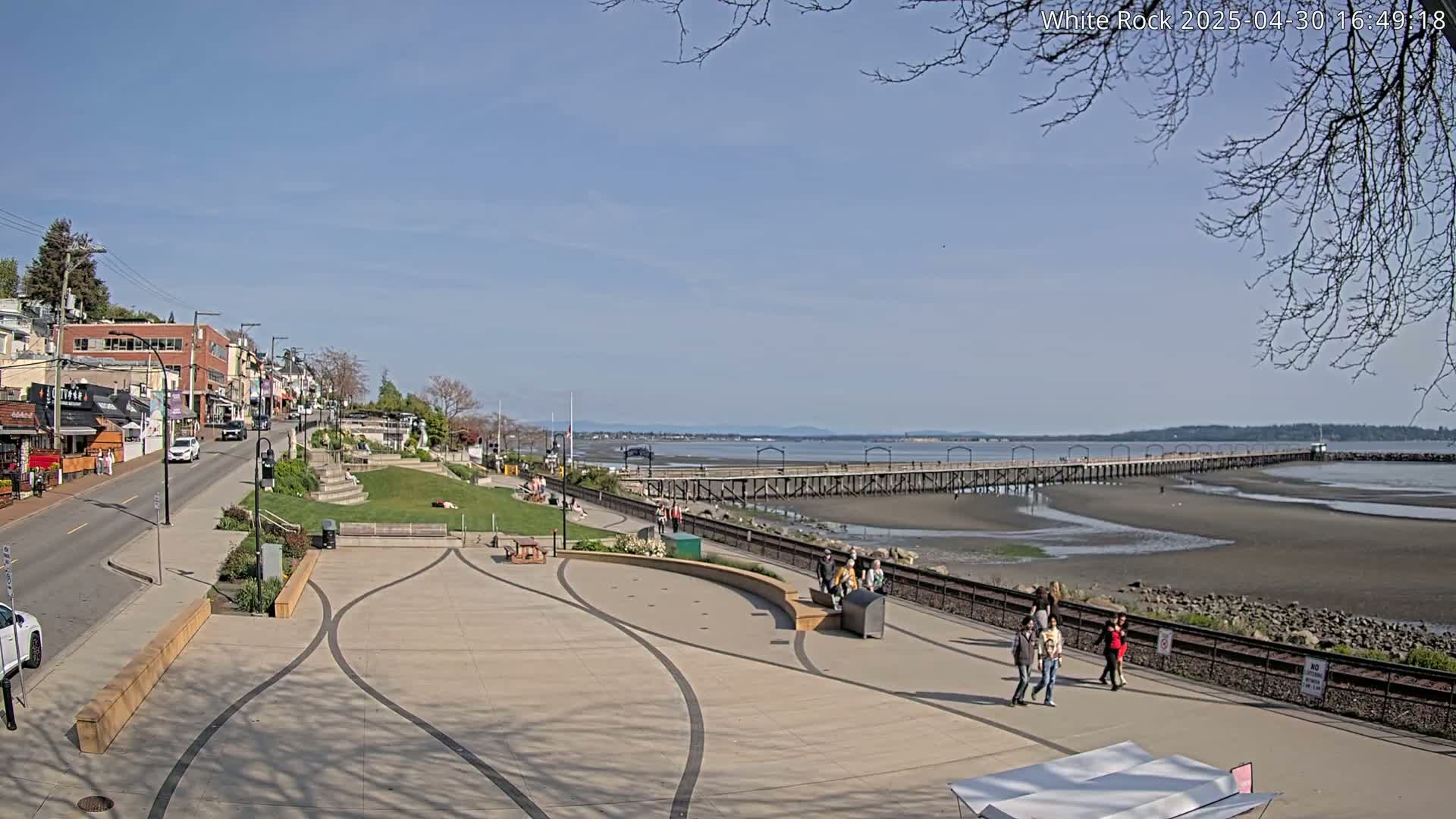 A paved waterfront plaza with a long pier extending into a tidal flat under a clear blue sky.