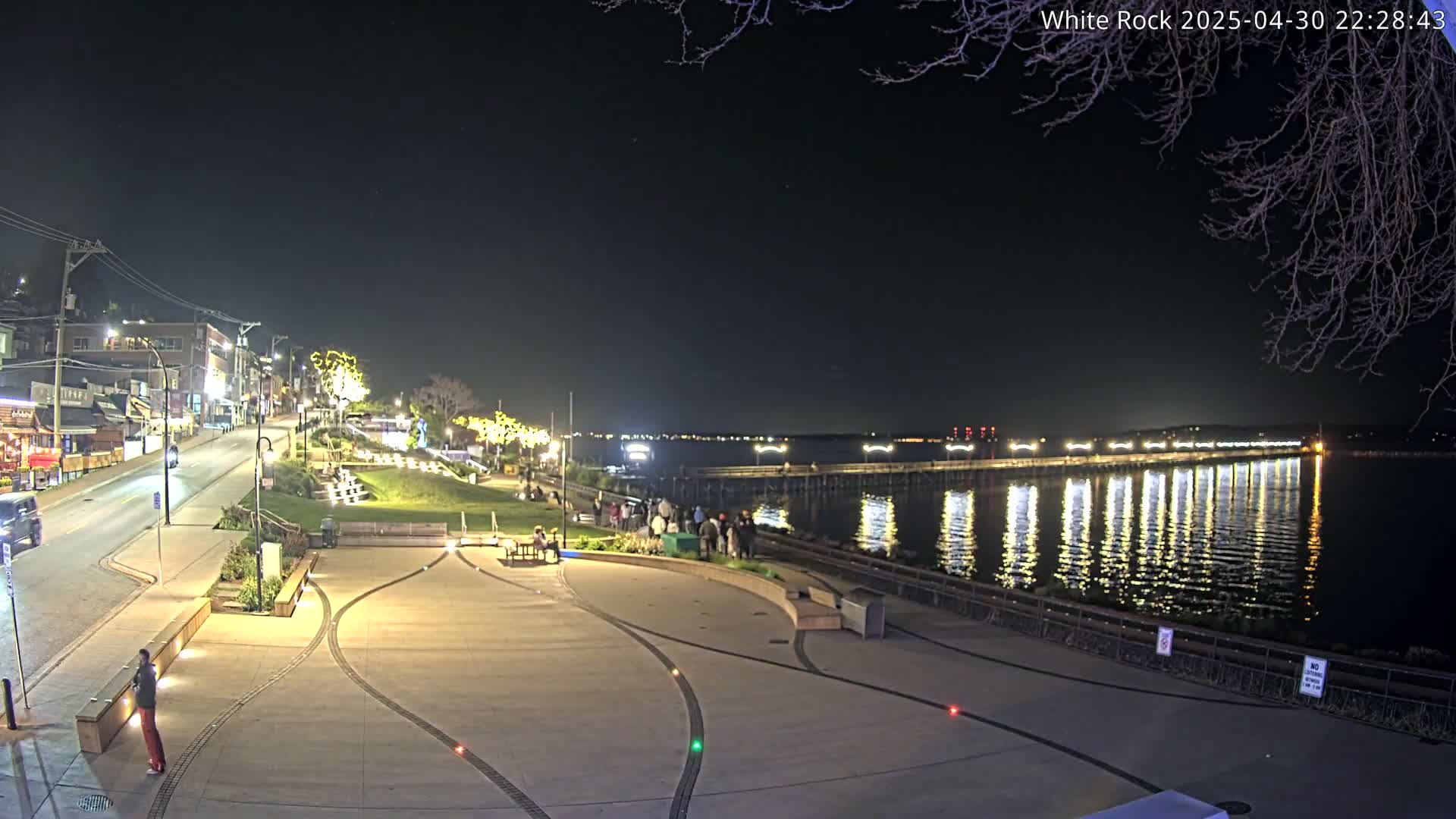 A nighttime view of a waterfront area with a long pier extending into calm water, illuminated by lights, and a paved plaza with people gathered nearby.