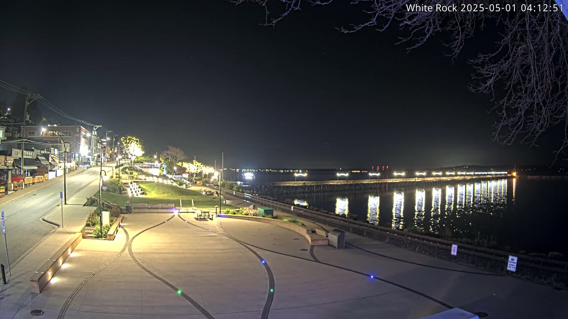 A nighttime view of a waterfront plaza with a paved area, benches, and landscaping, overlooking a calm body of water reflecting lights from a pier and distant shoreline.