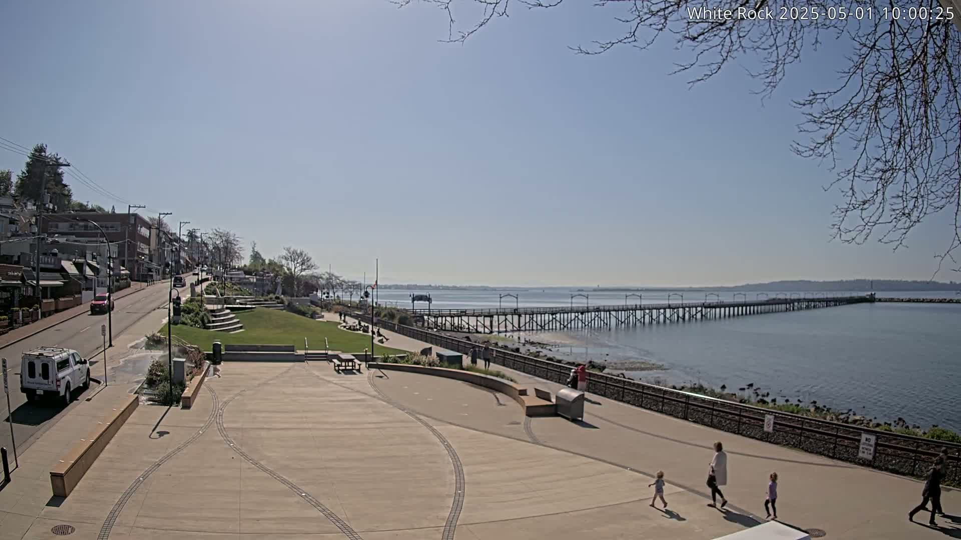 A paved waterfront plaza overlooks a long pier extending into a calm, blue bay under a clear, sunny sky.