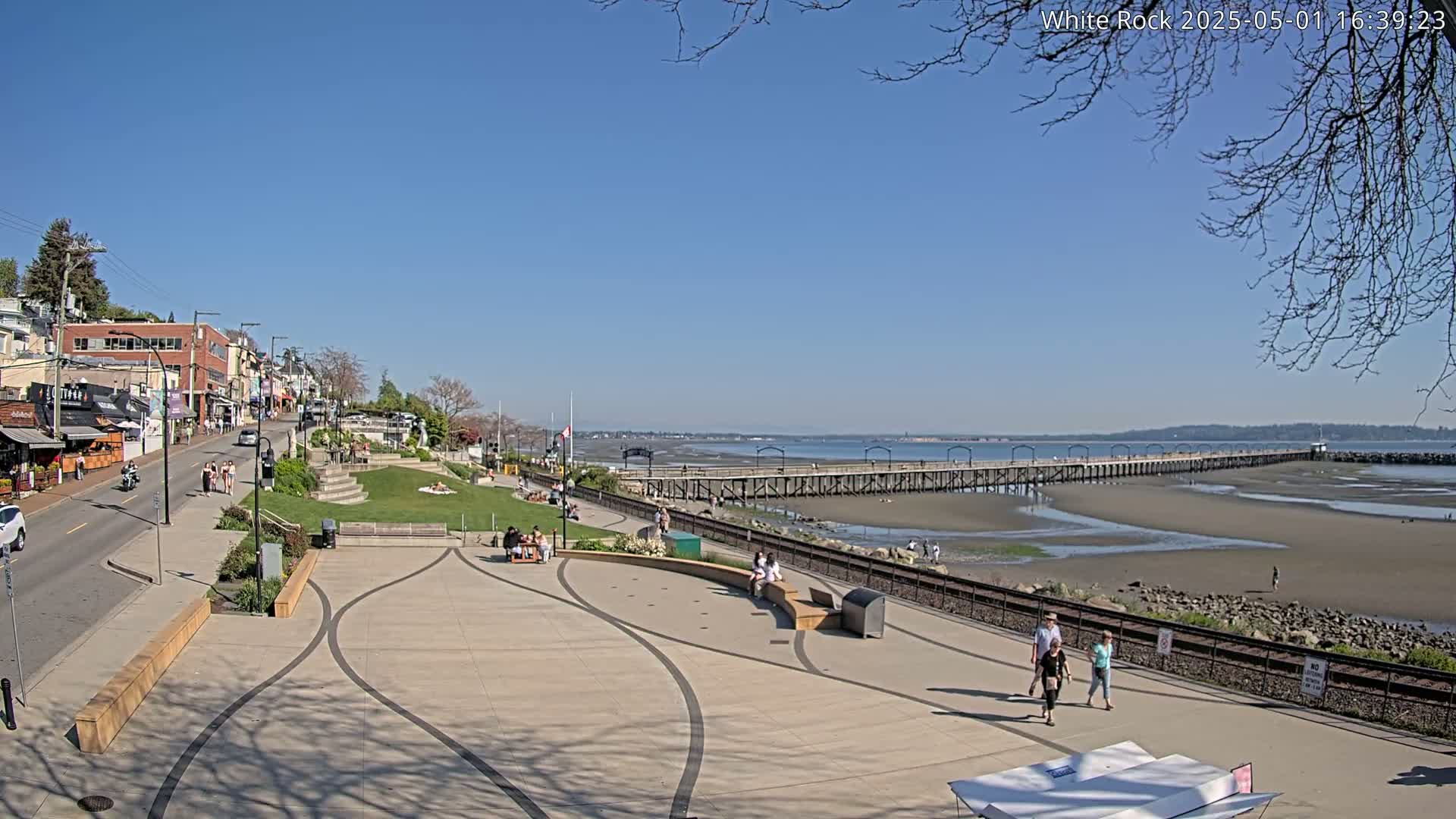 A sunny day reveals a waterfront town with a long pier extending into a tidal flat, people strolling along the paved promenade and nearby streets.