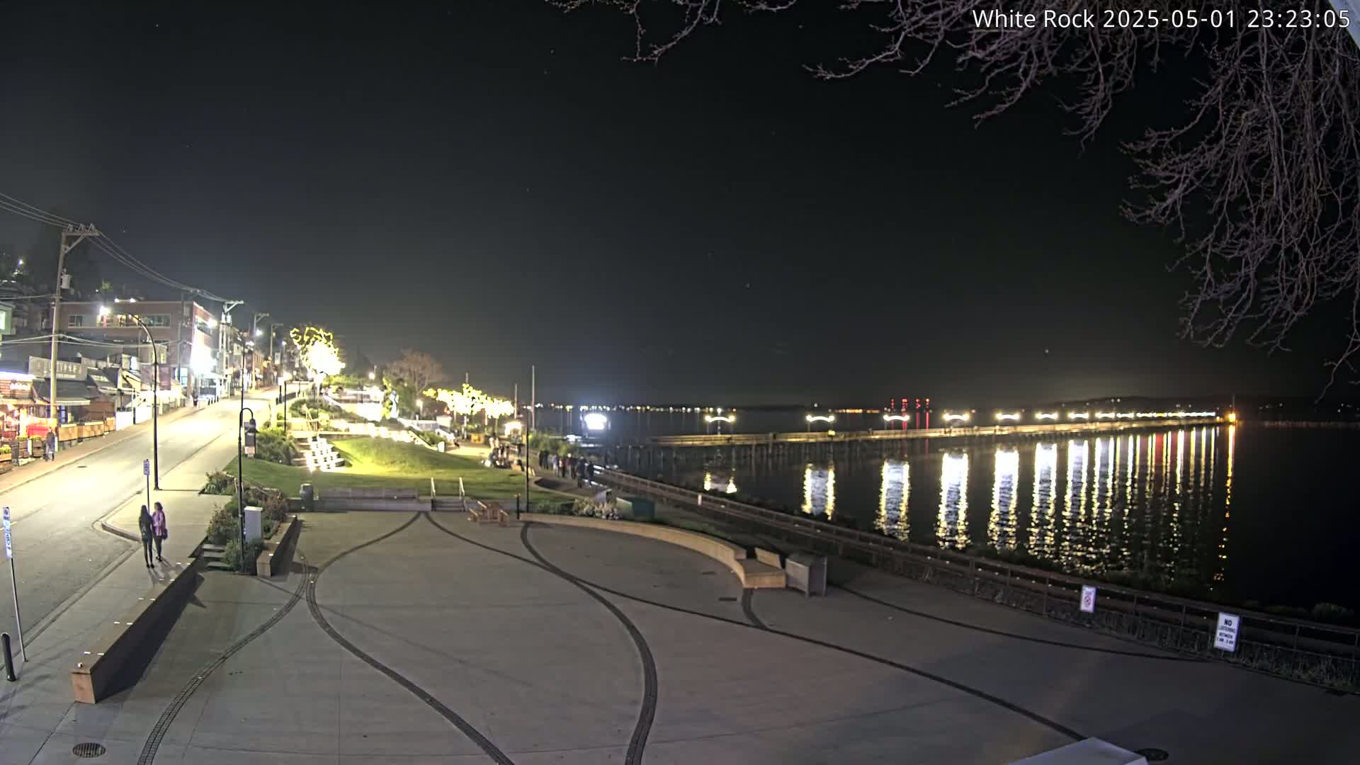 A nighttime view of a waterfront town with a long pier extending into calm water reflecting the lights, and a paved area with a few people walking.