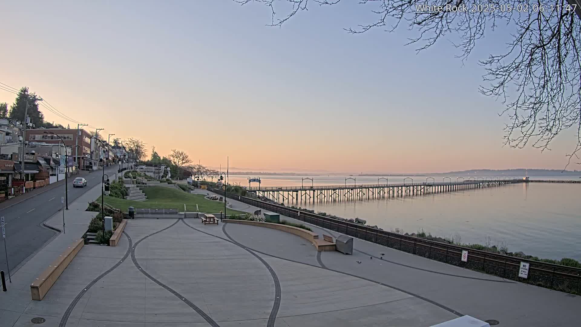 A long pier extends into calm water under a pale sunrise sky, next to a waterfront town with paved walkways and seating areas.