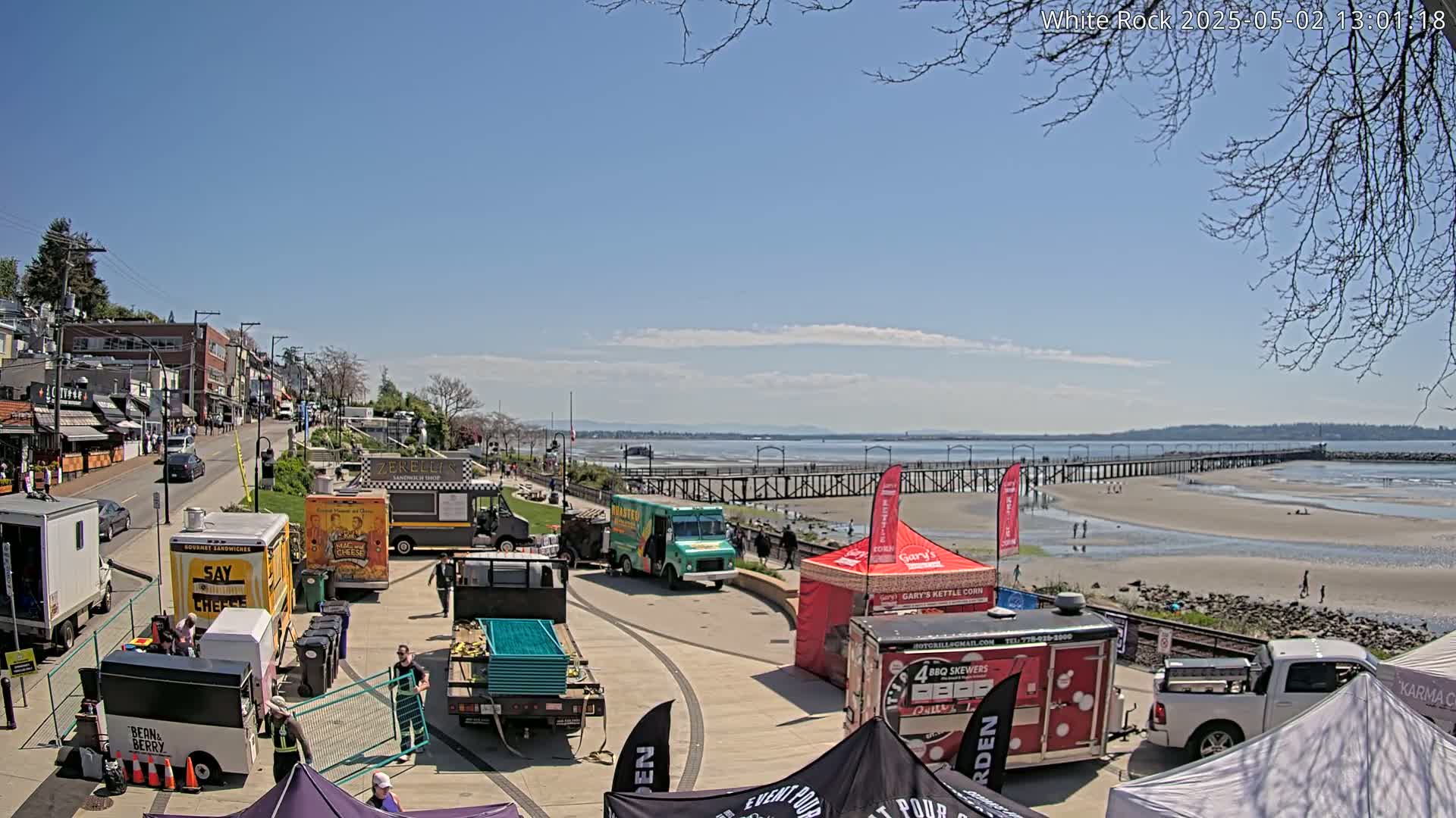 A sunny waterfront scene features numerous food trucks and vendor tents along a paved area, with a long pier extending into a sandy beach and calm water beyond.