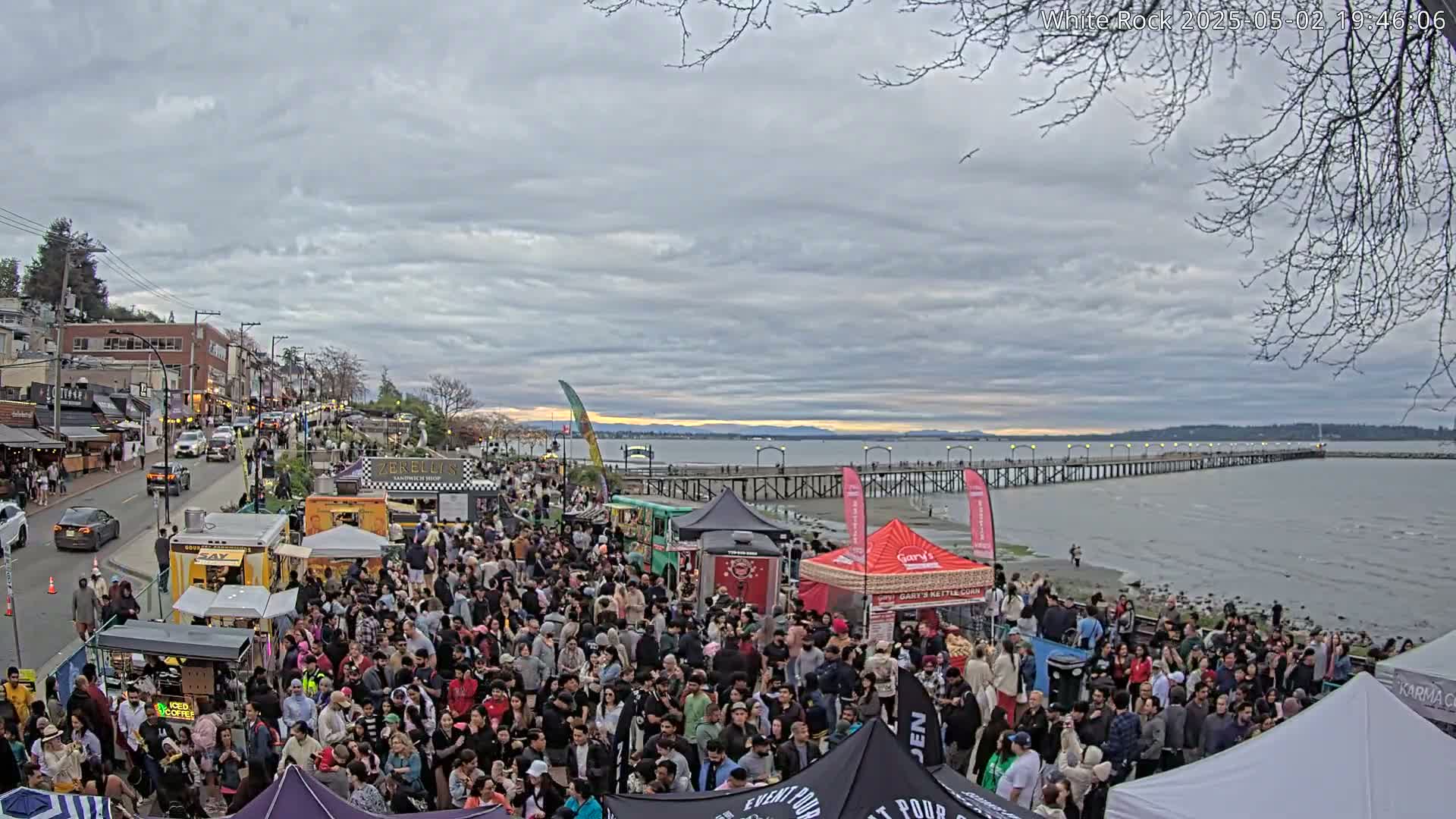 A large crowd gathers near a waterfront, featuring food trucks and tents under a cloudy sky, with a long pier extending into the calm water.