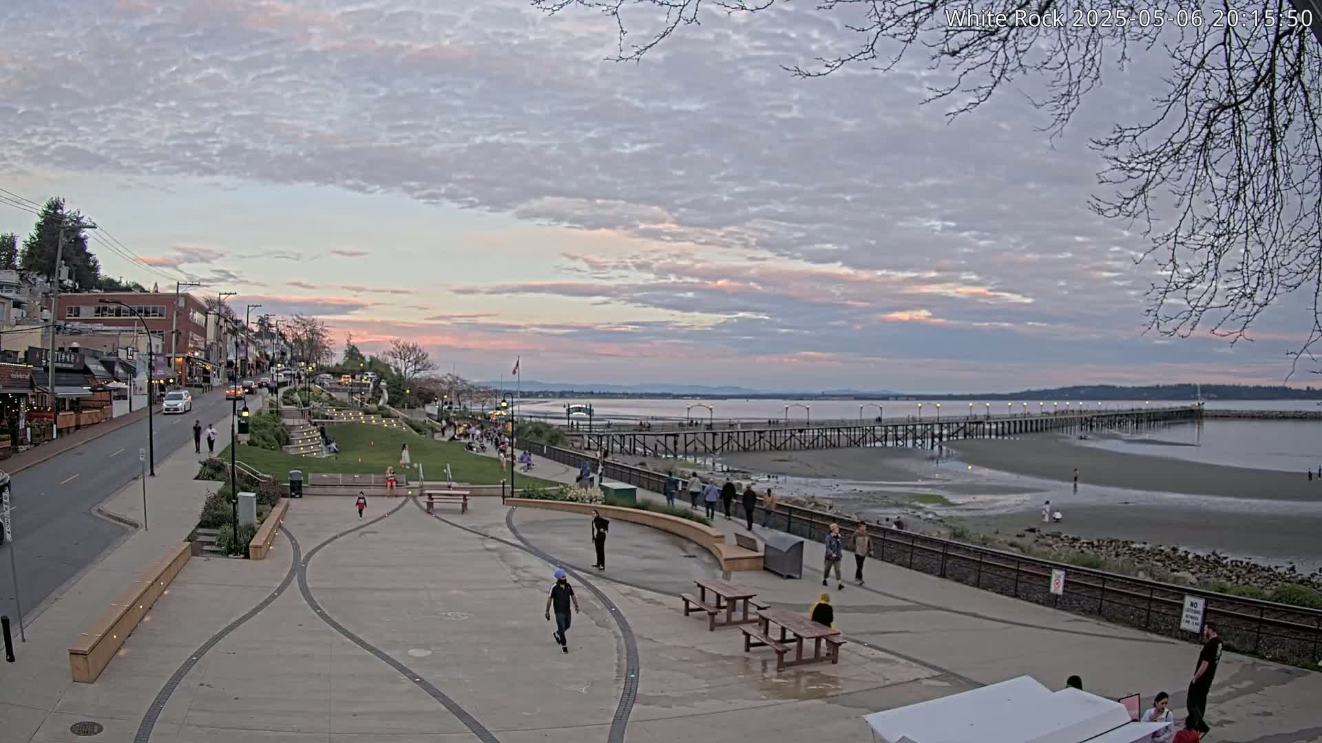 A seaside town square at dusk under a partly cloudy sky shows people strolling along a paved walkway toward a long pier extending into a calm bay.