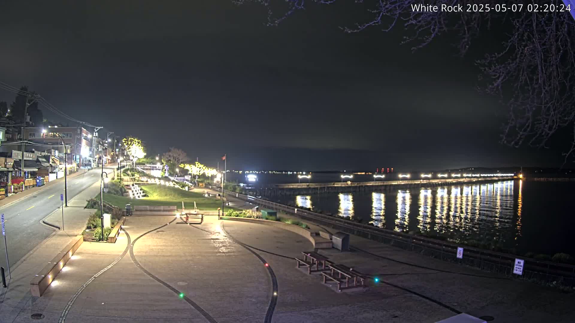 An empty, paved waterfront area at night is illuminated by lights, with a pier extending into the calm water reflecting the lights.