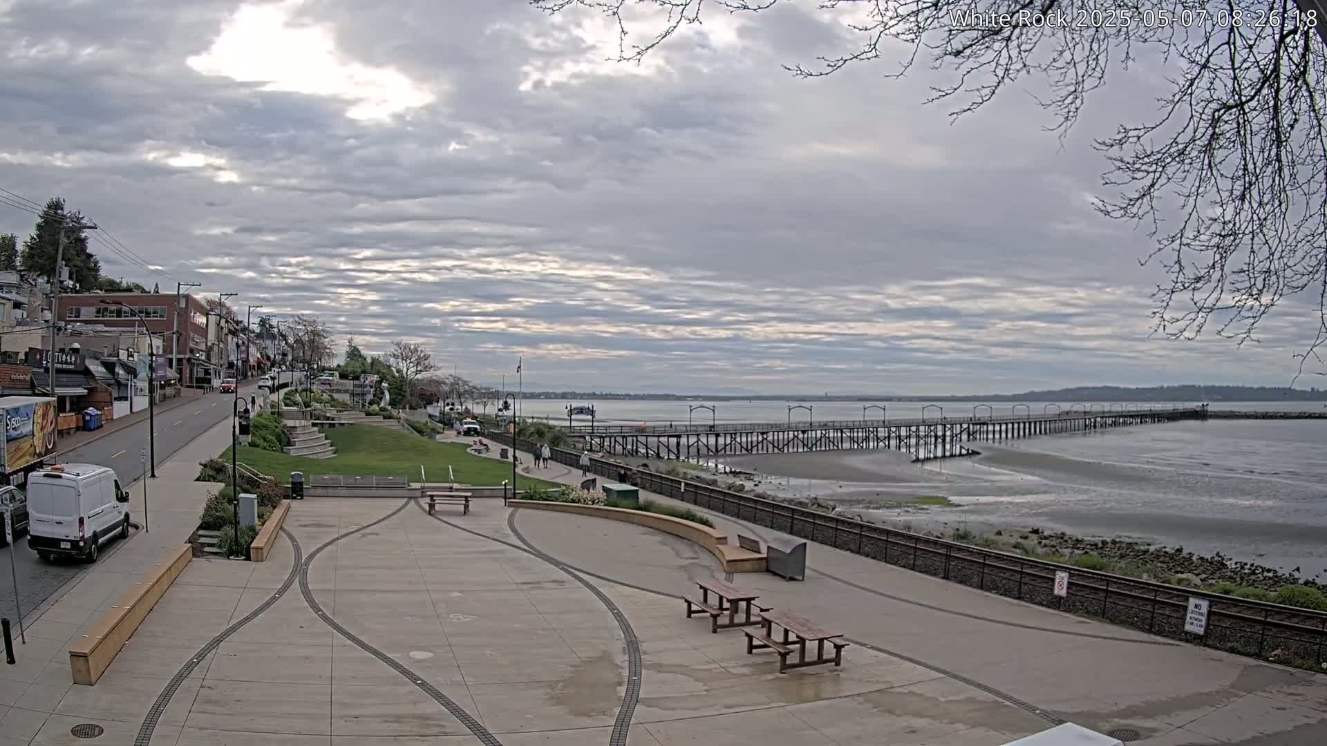 A paved waterfront plaza with picnic tables overlooks a long pier extending into a calm, grey-sky day over a body of water.