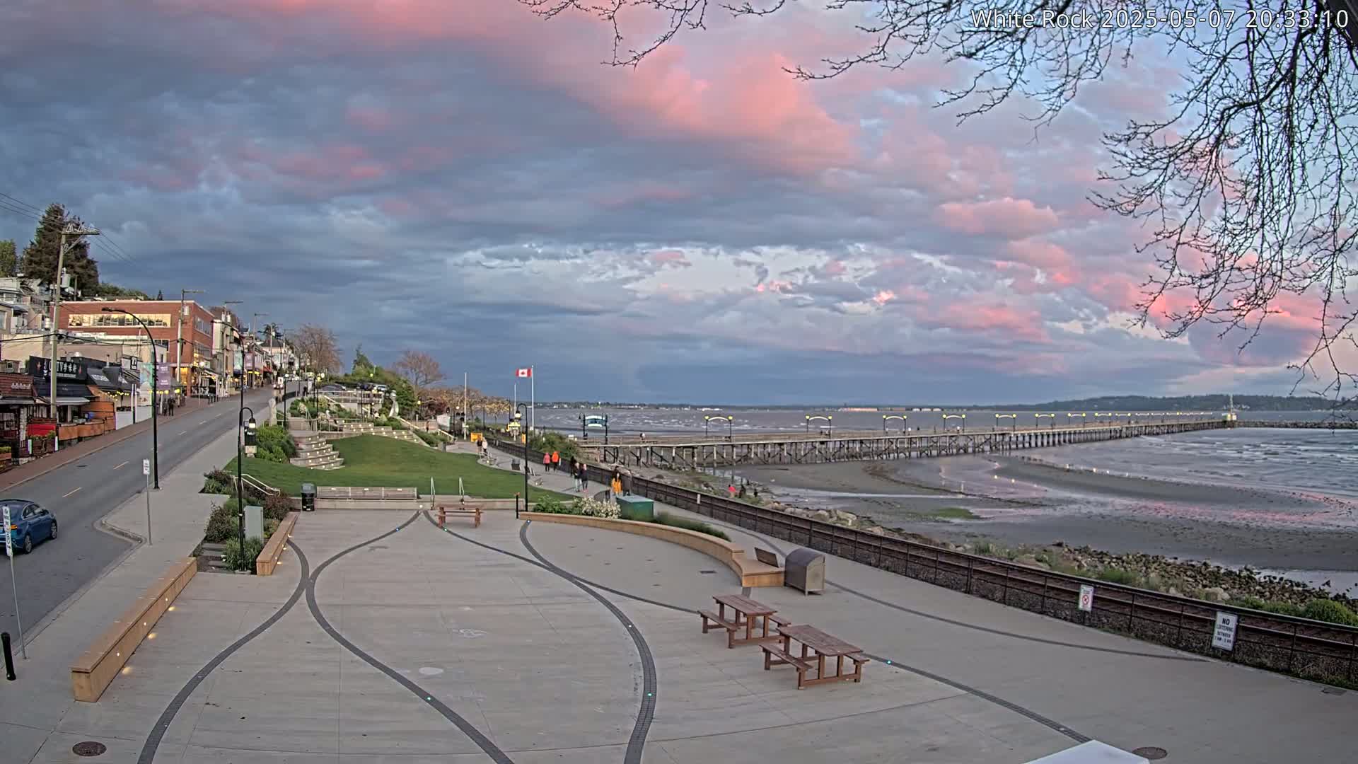 A long pier stretches into a calm bay under a partly cloudy sky with streaks of pink in the clouds, overlooking a paved waterfront area with benches and picnic tables near buildings and a road.