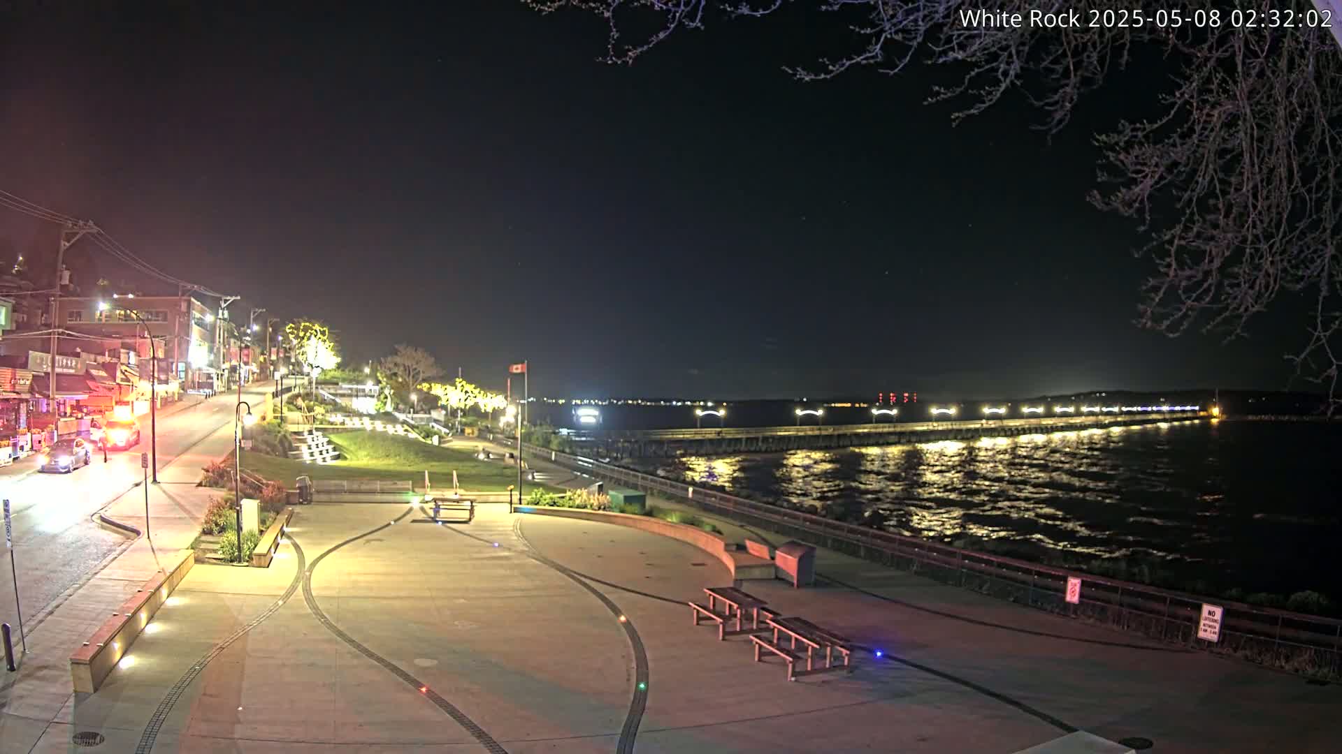 A nighttime view of a waterfront town square with a pier extending into a dark, slightly choppy body of water under a clear, starry sky.